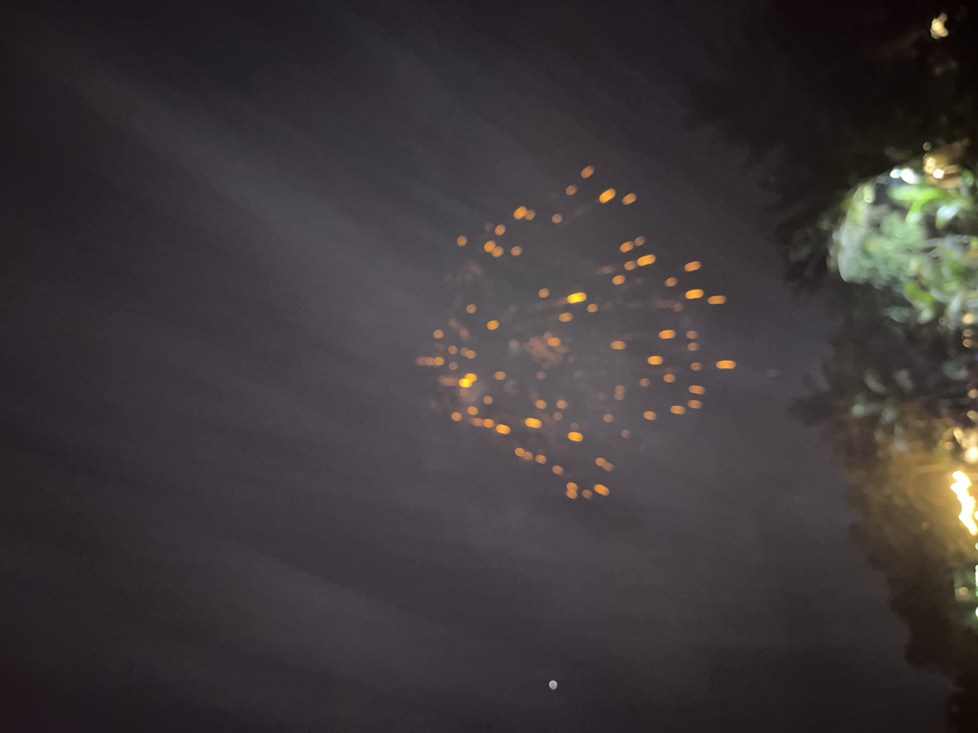 A nighttime scene in Habaraduwa, Sri Lanka, depicts a blurry display of what appears to be fireworks or sky lanterns against a dark, hazy sky. Numerous small, out-of-focus orange lights are scattered across the upper-middle portion of the frame, suggesting an event involving illumination in the sky. To the right, a blurred green mass with some bright white and yellow highlights indicates foliage, possibly trees, with some artificial lights, perhaps festive decorations, visible. The overall impression is one of a celebration or gathering taking place outdoors at night. The ambient light is dim, and the focus is soft, creating an atmospheric but indistinct view of the activity.