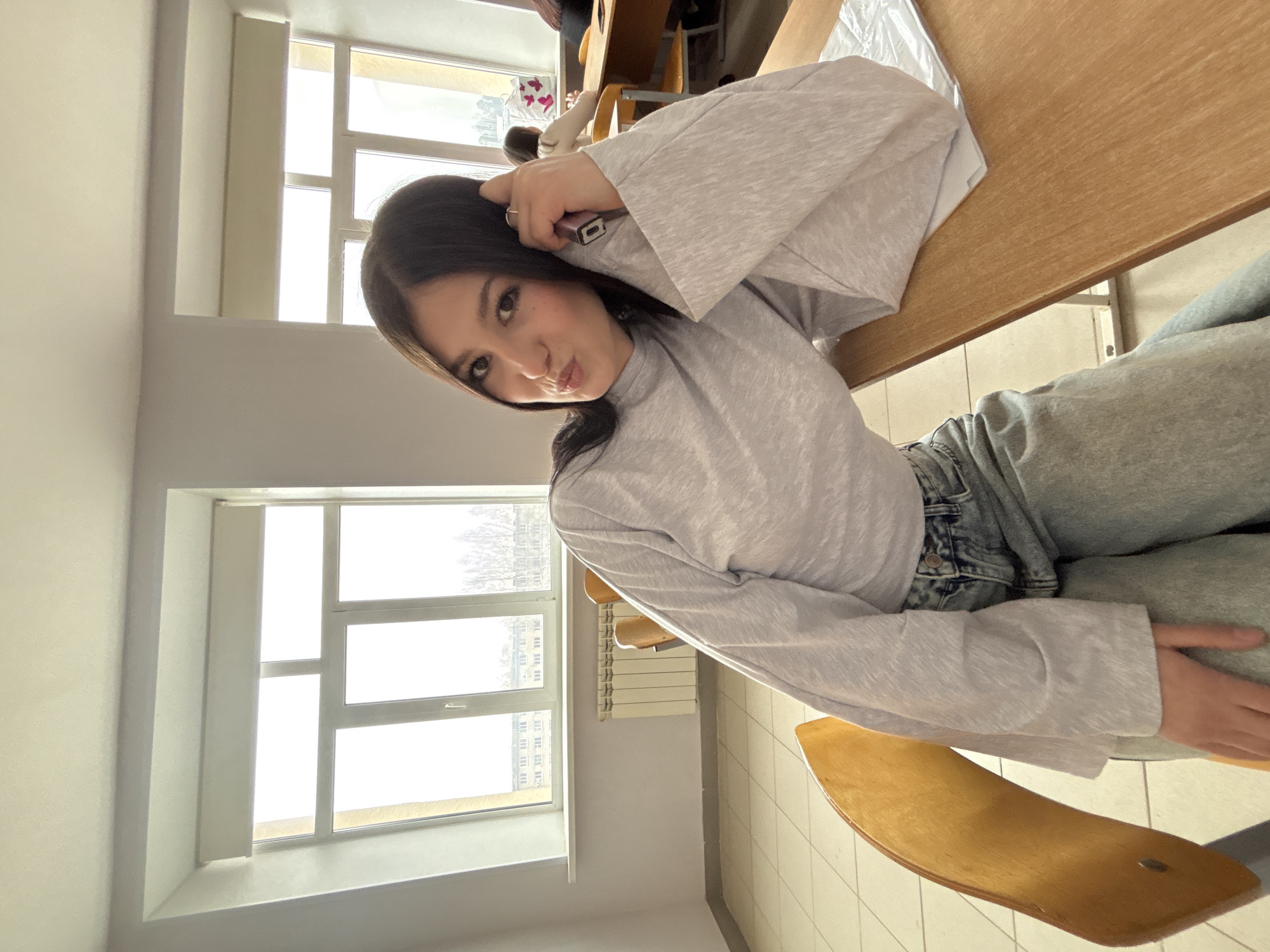 A young woman is posing for a selfie in a room with large windows, possibly a classroom. She is sitting at a desk, wearing a light gray, long-sleeved shirt and jeans. She is holding a phone to her head and making a face at the camera. The room has a radiator near the windows and there is another person in the background. Through the windows, buildings can be seen outside, and the architecture is consistent with that found in Chaikovsky, Russia.