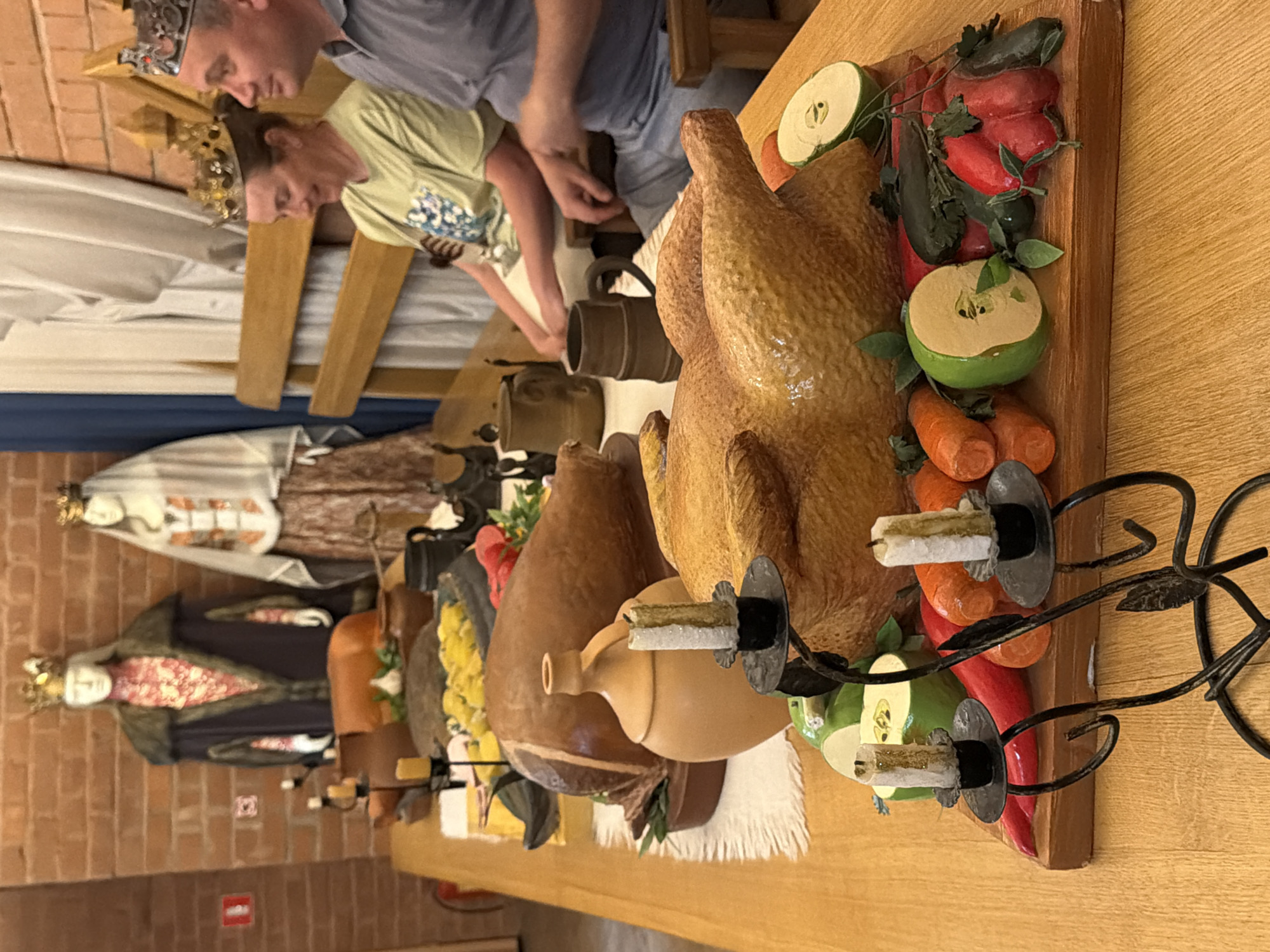 In the city of Unknown, Unknown, a long wooden table is set for a medieval feast. The table is laden with various food items, including a large roasted turkey, a whole roasted pig, and a platter of yellow fruit. A ceramic jug and several earthenware mugs are also present. In the background, two mannequins dressed in regal attire, complete with crowns, stand as if presiding over the feast. Two people, a woman and a man, both wearing crowns, are seated at the table, appearing to be guests or participants in the event. The atmosphere suggests a historical reenactment or a themed celebration.