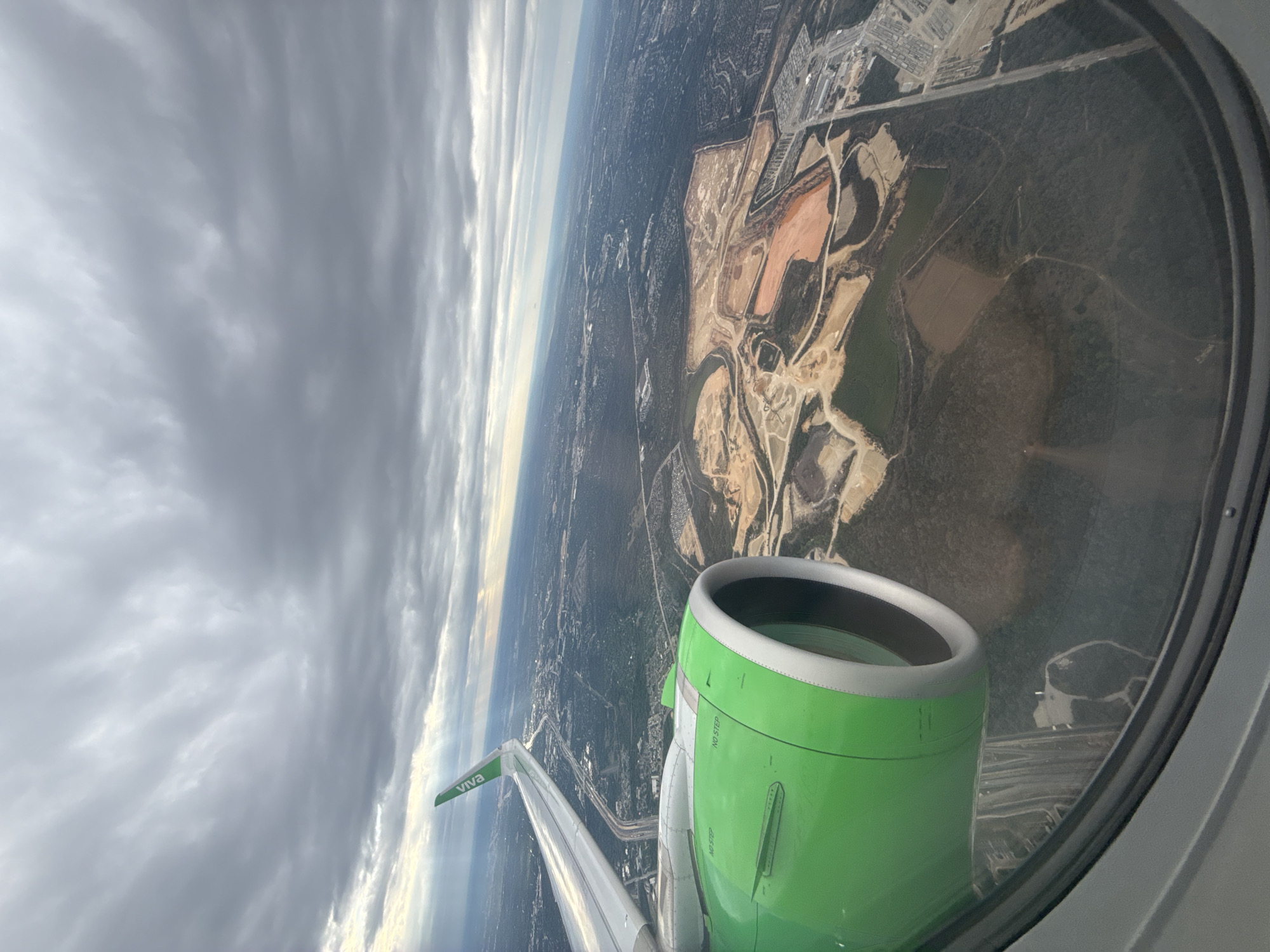 The image shows an aerial view from inside an airplane, likely during descent or ascent. Outside the window, there's a green engine of a Viva Aerobus plane and a wing with the "Viva" logo. The landscape below features a mix of urban and natural environments, including what appears to be an open pit mine, with red soil contrasting with the green vegetation. The urban area shows buildings and roads, including highways and clusters of buildings. The sky is covered with gray, overcast clouds, suggesting a cloudy day. The location is possibly near San Antonio, United States of America, based on the landscape's characteristics and presence of large-scale open pit mining.