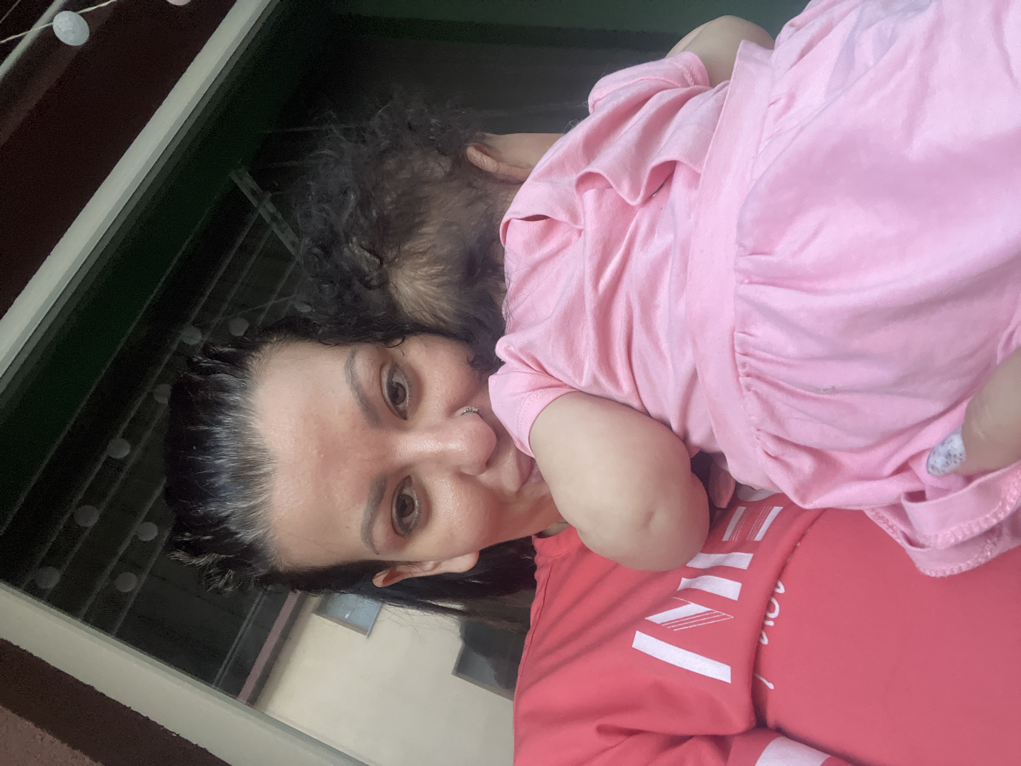 A woman with dark hair and a nose ring is holding a baby. The woman is wearing a coral-colored shirt with white lettering. The baby is wearing a pink dress and is facing away from the camera, with its head tucked into the woman's shoulder. They are standing in front of a dark green door with some string lights visible above. The scene appears to be in an outdoor setting, possibly a balcony or patio, with part of a building visible in the background. The image is a selfie taken by the woman.