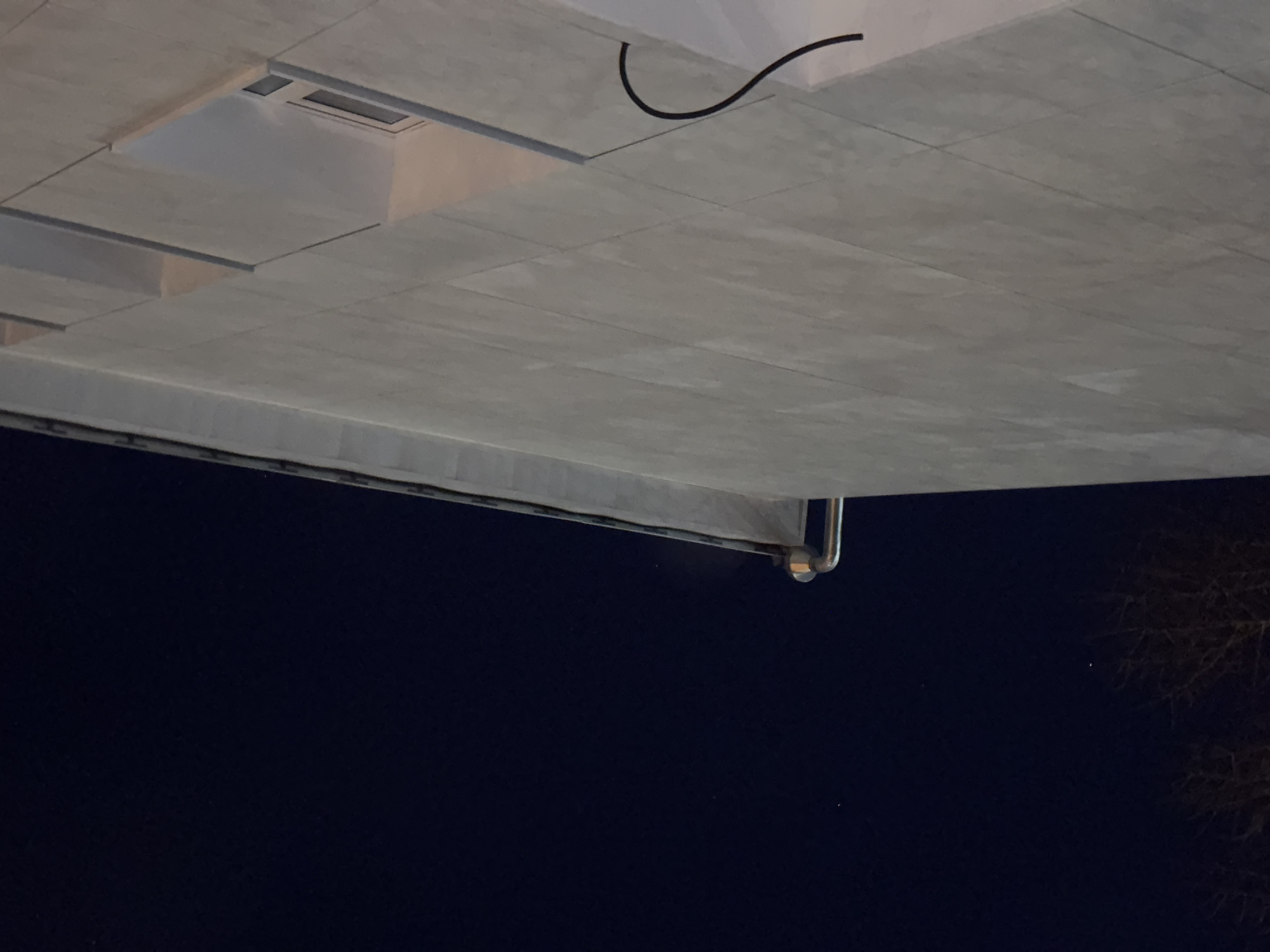 A low-angle, nighttime shot shows a gray, tiled ceiling with what appears to be a recessed light fixture in the upper left. A black cable runs along the surface of the ceiling. In the lower portion of the image, below the ceiling, is a dark area suggesting night sky. At the bottom edge, visible branches of a tree are present on the right side of the image. In the lower-left quadrant, a gutter or pipe end is visible. The location may be in Perm, Russia, but this cannot be verified from the image.