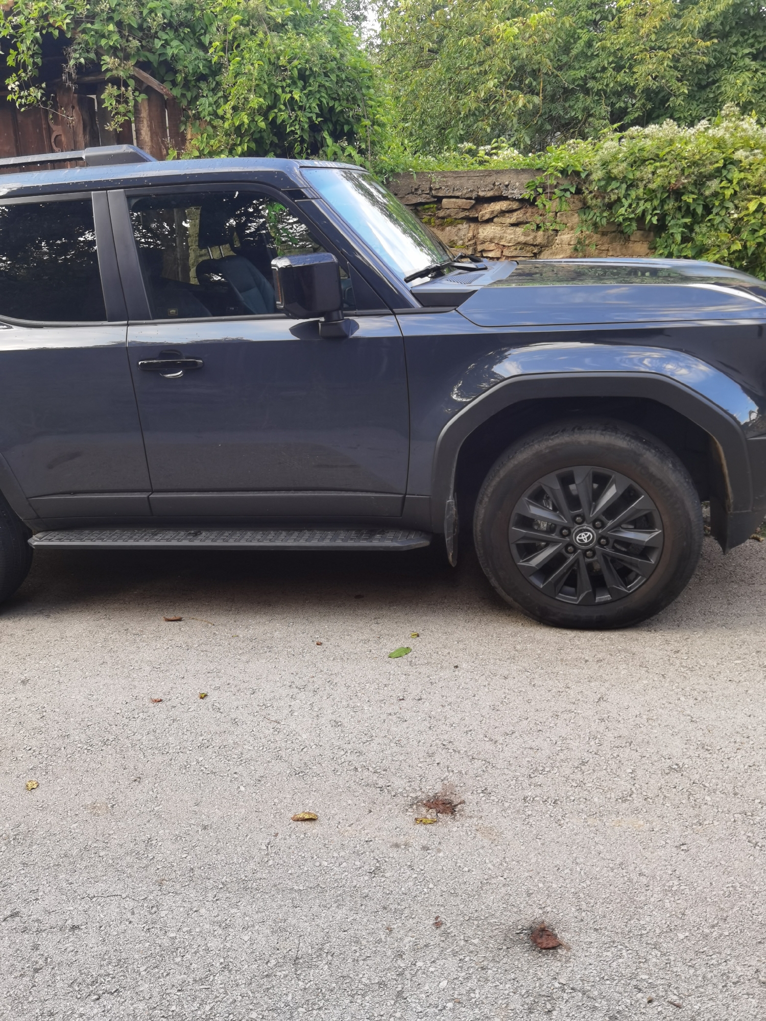 The image shows the side view of a dark gray SUV parked on an asphalt surface. The vehicle has a running board, black rims with a Toyota logo, and tinted windows. Behind the vehicle, there is a stone wall with dense green foliage growing over it, and further back, a wooden structure is visible, partially covered by plants. The ground is a rough asphalt with some scattered leaves and what appear to be small, dark stains. The scene suggests an outdoor location, possibly in a suburban or rural area, consistent with the village of Lomets, Bulgaria, given the rustic background elements.