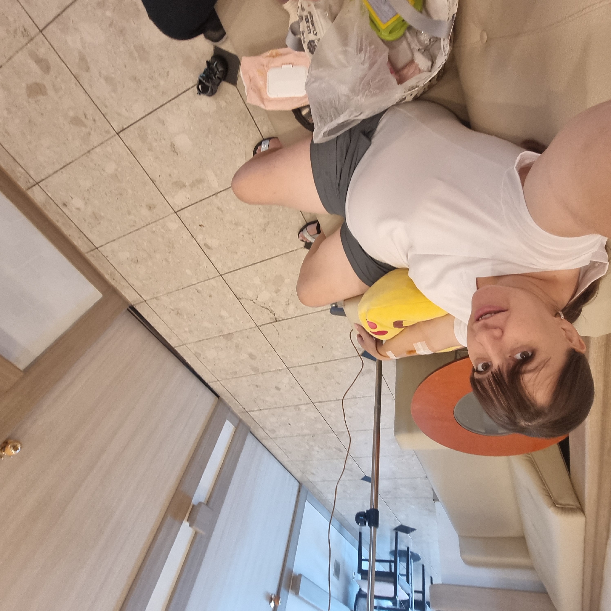 A woman with dark brown hair and bangs is sitting on a beige couch in what appears to be a waiting room or hospital setting. She is wearing a white t-shirt and grey shorts. An IV drip is attached to her left arm, which is resting on a yellow emoji-shaped pillow. The floor is tiled with a beige and cream pattern. In the foreground, on the floor, are various items including a plastic bag, a pink package of wipes, and a pair of dark shoes. A metal IV stand is positioned behind her. The room has light-colored walls and doors, with a hallway visible in the background. There is a circular wooden table with a dark center to the left of the woman. The overall impression is that of a medical facility. The location is not specified to be in Wonju-si, South Korea.