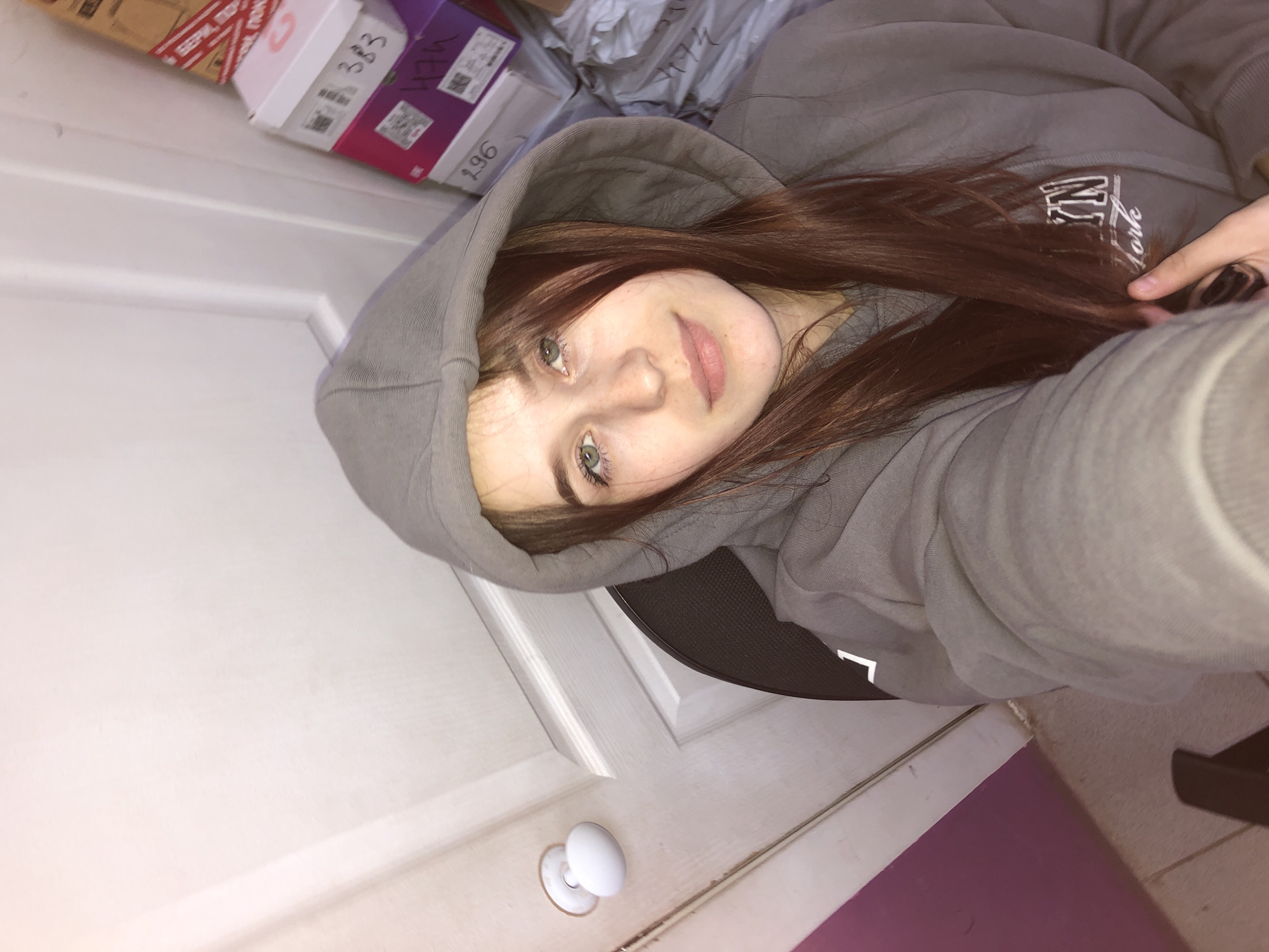 A young female, appearing to be in the city of Фоки, Russia, is depicted in a close-up selfie-style photograph. She has light skin, green eyes, and long reddish-brown hair, which falls over her shoulders and chest. She is wearing a grey hooded sweatshirt with partially visible white text "BRKLYN New York" on the front. Her right hand, holding the camera, is visible at the bottom right. She is seated, with a portion of a dark-colored chair back visible behind her left shoulder. The background features a white paneled door with a round white doorknob on the left. To the right of the subject, a stack of various-sized boxes is visible, predominantly white and purple/pink, with handwritten numbers such as "383", "474", and "196" on their sides, suggesting storage or inventory. A large cardboard box with red and white tape bearing Cyrillic text is positioned at the top of the stack. Below the boxes, crumpled light-colored material is visible. The lower left corner shows a segment of a light-colored tiled floor next to a dark pink/purple wall section.