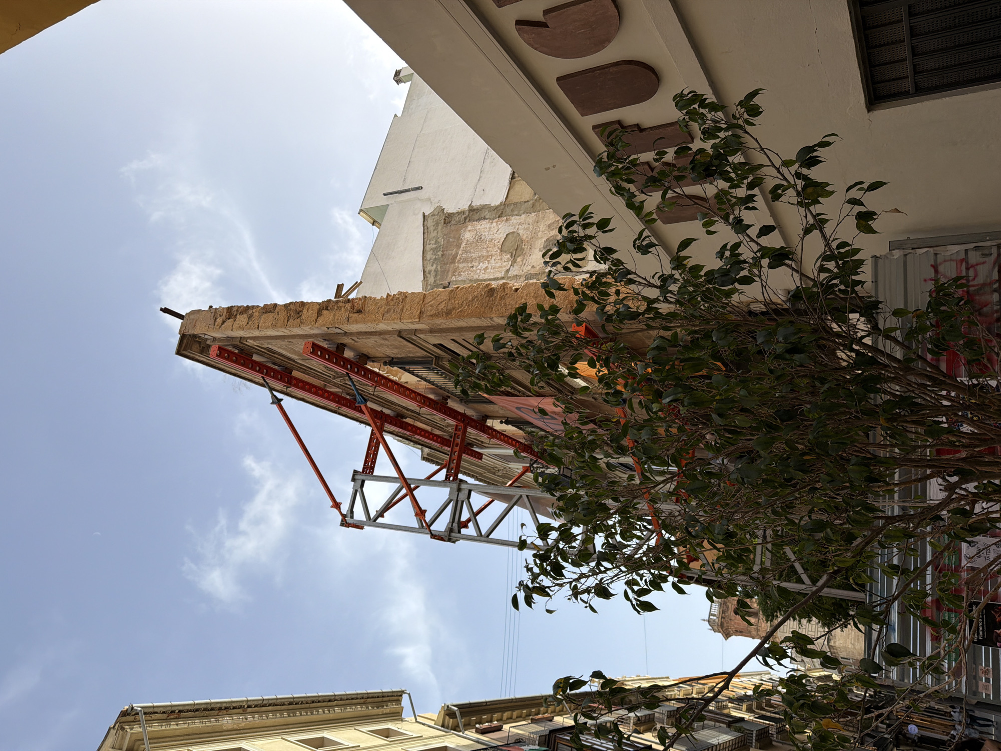 A low-angle, outdoor shot shows the side of a crumbling building undergoing repairs. A complex metal structure, painted red and silver, is braced against the building's damaged facade. The building itself appears to be a brick structure with sections missing, revealing exposed interior walls. To the left and right of the damaged building are other adjacent buildings. In the foreground, a bushy green tree with bare branches partially obscures the view. The sky above is a clear blue with a few wispy clouds. A streetlamp and some signage are visible in the distance. The overall scene suggests an urban environment with ongoing construction or demolition work.
