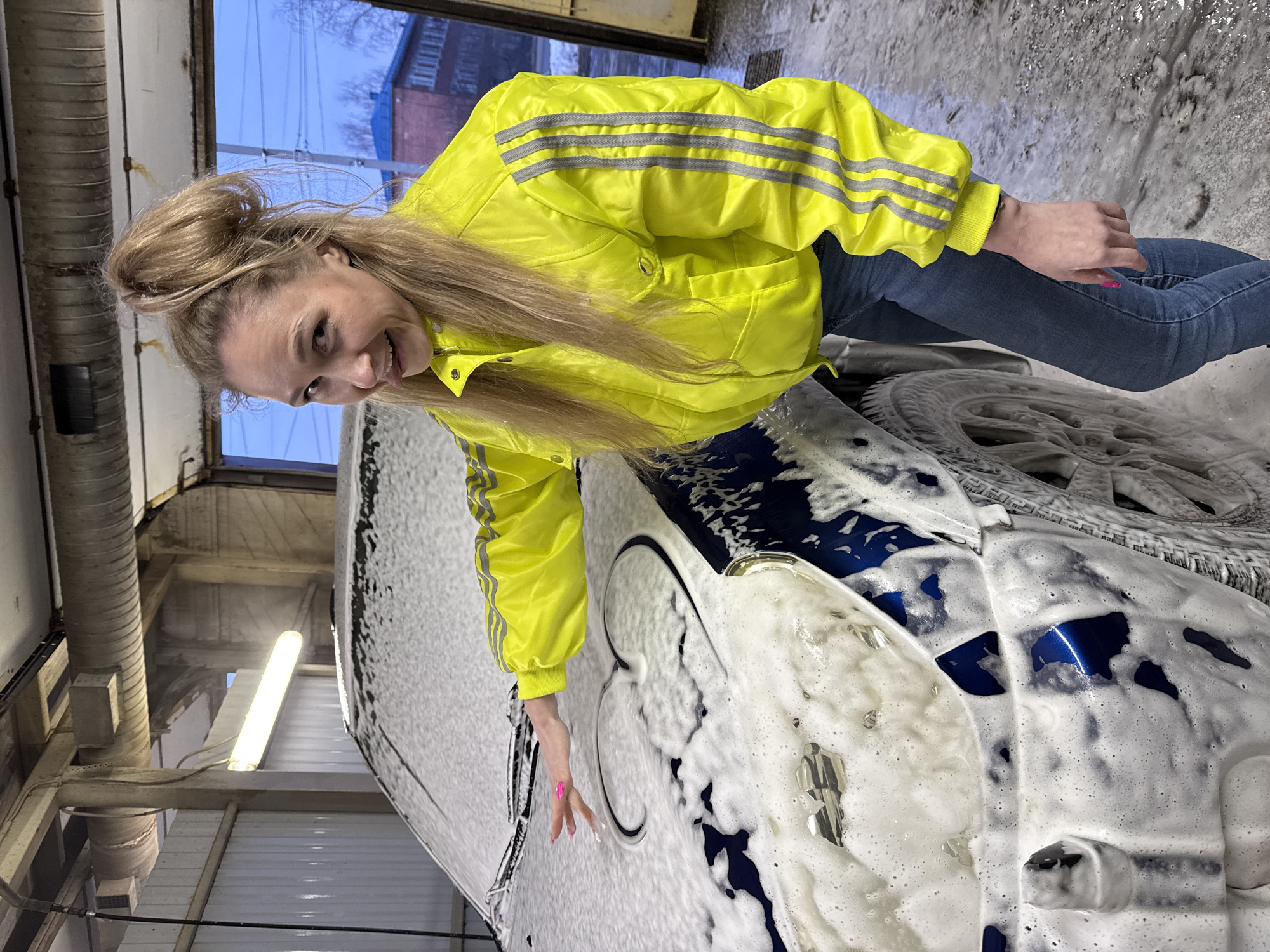 An indoor scene depicts a female subject posing next to a blue car covered in white cleaning foam within a car wash bay in Chaikovsky, Russia. The woman, with blonde hair styled in a high bun and loose strands, wears a vibrant yellow jacket featuring three grey stripes on each sleeve, paired with blue denim jeans. Her right hand, exhibiting pink nail polish, is positioned on the car's foamy hood, where a heart shape has been drawn or outlined in the foam. She looks directly at the camera with a playful expression, her tongue slightly extended. The vehicle's hood partially reveals a Chevrolet emblem beneath the foam. The immediate environment includes a wet, concrete floor, corrugated wall panels, and exposed overhead fluorescent light fixtures. Beyond a large window in the background, a blue sky, suggestive of late afternoon or dusk, is visible above distant trees and a multi-story brick building with a blue roof.