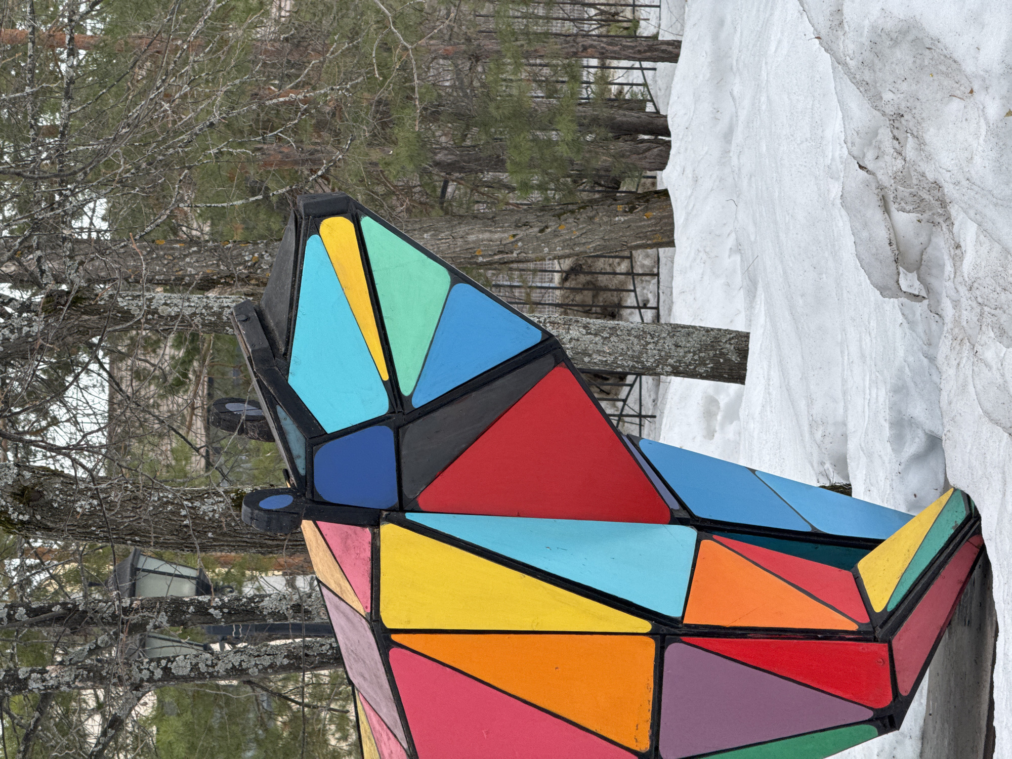 The image captures a vibrant, geometric sculpture situated in a snowy outdoor setting, likely in a park or public space. The sculpture is constructed from triangular and rectangular segments painted in a variety of bright colors, including shades of blue, green, yellow, orange, red, and purple. These colorful segments are framed by black edges, creating a striking contrast.
In the background, there are bare trees with intricate branches, suggesting a late winter or early spring season. A patch of snow is visible to the right of the sculpture, indicating cold weather conditions. There's a hint of a fence or railing in the distance.
Based on the prompt's reference to Chaikovsky, Russia, it is reasonable to assume that this is the location of the scene. However, without additional information, I cannot provide more precise details about the exact location or any people, actions, or interactions.