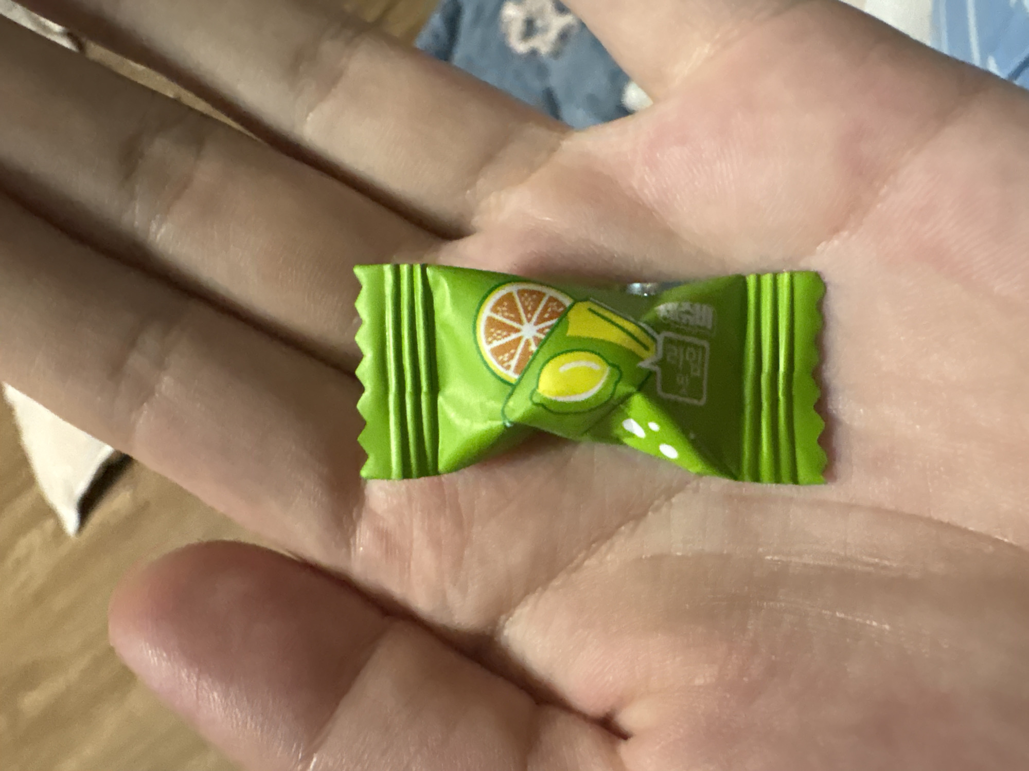 An extreme close-up shot shows a person's hand holding a green, individually wrapped candy. The candy wrapper has a serrated edge at the top and bottom and features an illustration of a sliced orange and a lime. Text in an East Asian script is visible on the lower portion of the wrapper. The hand is open, with the candy resting in the palm, and the fingers are curled slightly. The background is blurred, showing a textured surface and a hint of fabric with a pattern. The lighting appears to be artificial and overhead, casting subtle shadows on the hand.