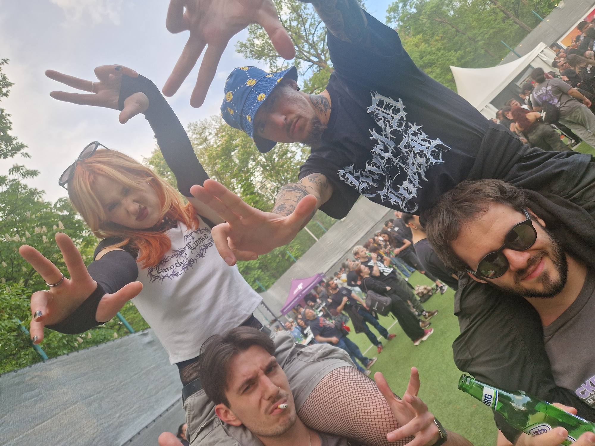 A group of four people poses for a photo at an outdoor event, possibly a music festival, judging by the crowd in the background and the presence of a tent. One person is sitting on the shoulders of another, while a third person is sitting on the shoulders of a fourth. All four individuals are making hand gestures towards the camera, seemingly posing for the photo. The environment appears to be a park or grassy area, with trees visible in the background. The overall atmosphere seems festive and casual. The city and specific location are unknown.