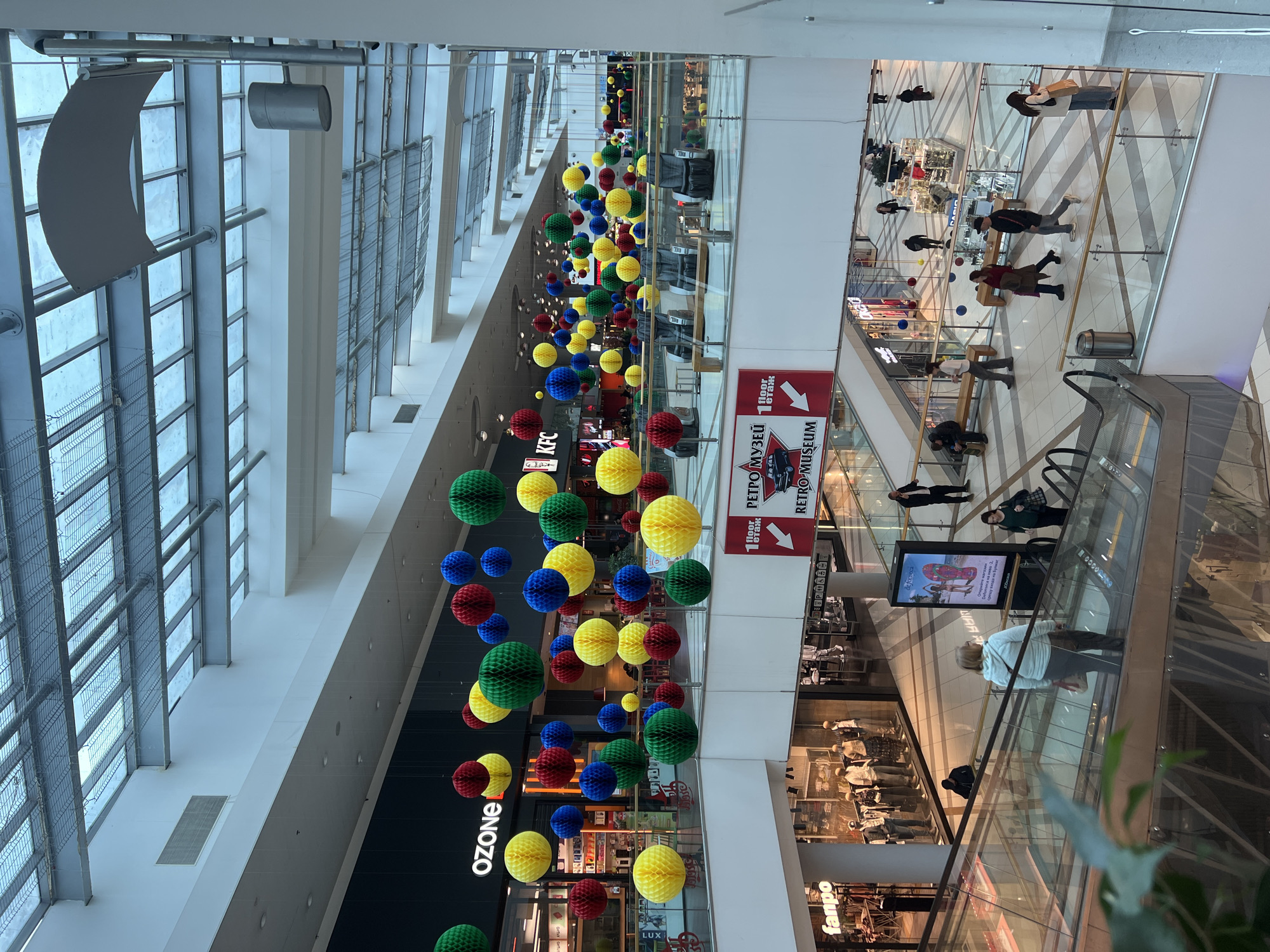 The image depicts an interior view of Grand Mall Varna, Bulgaria, focusing on a multi-story atrium. The overhead structure features a large, transparent glass skylight supported by white metal beams and gridded elements. Numerous spherical, honeycomb-style paper lanterns in red, yellow, green, and blue are suspended at various heights throughout the central space.
On the upper visible level, storefronts include "OZONE" and "KFC." Five dark grey massage chairs are arranged beyond a glass railing, accompanied by two light-colored benches.
The mid-level features a glass and metal railing from which a prominent red and white banner hangs. The banner displays "1 floor," "РЕТРО МУЗЕЙ" (Retro Museum), and "RETRO MUSEUM" with an arrow pointing to the left. Below this, a "fanpo" retail store is visible, showcasing mannequins behind a glass facade.
Multiple individuals are present across various levels. In the mid-foreground, a person with light hair, wearing a light-colored top and dark pants, is descending an escalator while carrying a shoulder bag. Further right, a person in a dark green top and dark pants, holding a dark handbag, ascends an escalator.
On the lowest visible floor, a person in a dark jacket is seated on a light-colored wooden bench. Another individual in a white long-sleeve shirt and dark pants walks past the bench. A woman in a maroon coat and a man in a black jacket and jeans walk in opposite directions across the tiled floor. Another woman in a white top and dark pants, carrying a white shoulder bag, looks at a mobile device while walking. In the background, other individuals are scattered, some near retail kiosks, including one marked "CASIO." A man in a dark jacket stands near a pillar, speaking on a mobile phone. A vertical digital display screen advertises "De Fonseca" footwear.
The flooring consists of light-colored tiles, some with dark geometric patterns. Silver cylindrical trash receptacles are positioned near the escalators. A blurred green plant occupies the bottom left foreground.