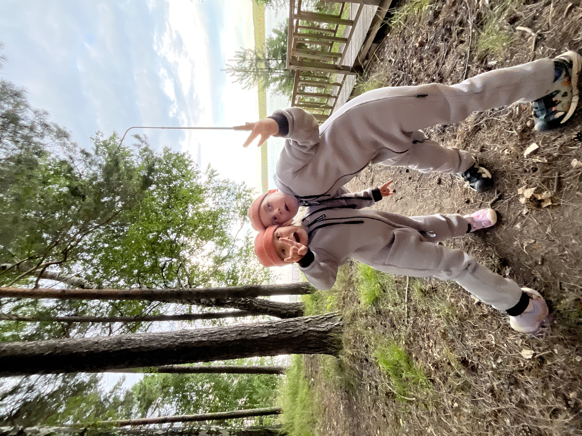 Two children, one slightly behind the other, are posing for a photo in an outdoor setting. Both are wearing light grey hooded jumpsuits and orange beanies. The child in front is making a peace sign with their left hand and their mouth is open as if speaking or laughing. The child behind them has their left arm raised, also making a peace sign with their fingers. They are standing on a path covered in dry leaves and grass, with trees on either side. In the background, there is a wooden boardwalk leading to a body of water, likely a lake, with a slightly overcast sky. The image appears to be taken during the daytime, possibly late afternoon given the lighting. The overall mood seems playful and energetic. The image is taken from a slightly upward angle, looking towards the sky. There is a thin, metallic pole extending into the frame from the left side.
