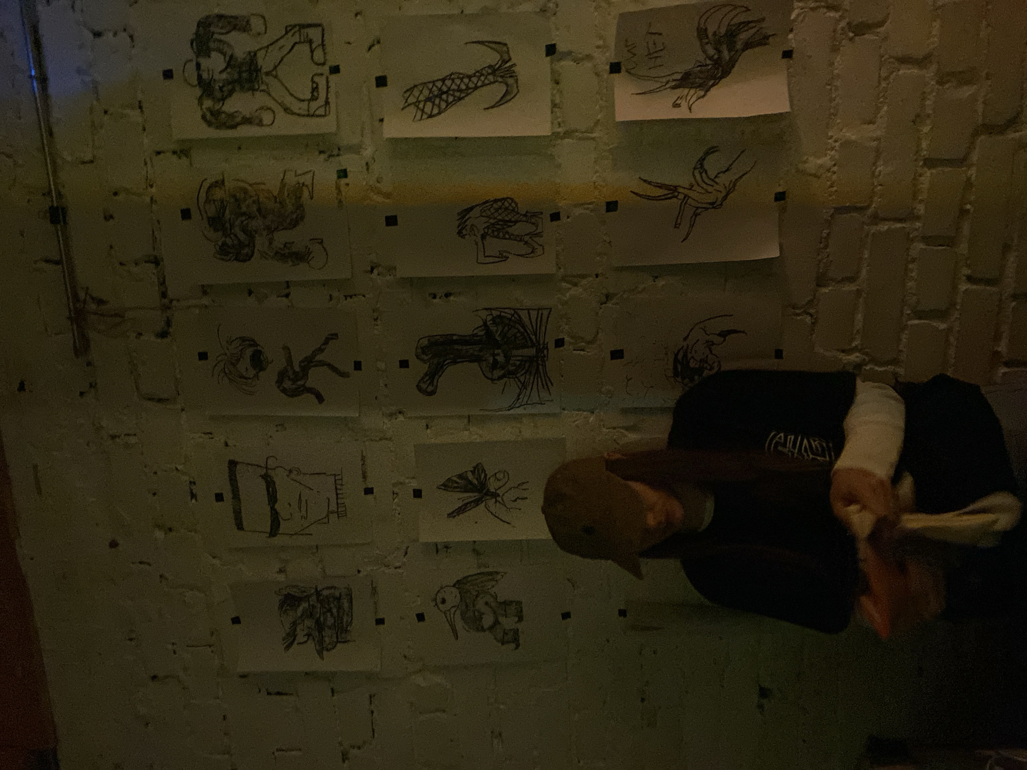 A low-angle shot inside what appears to be a brick-walled room with ten drawings or sketches pinned to the wall. The drawings include depictions of figures and creatures. A person wearing a dark jacket, a light-colored long-sleeve undershirt, and a cap is partially visible, possibly seated on a chair, seemingly obscured from the wall's view. The lighting in the room is dim, and there is limited visibility of the other objects and the person's facial features. The city of Moscow, Russia, is not discernible in this image.