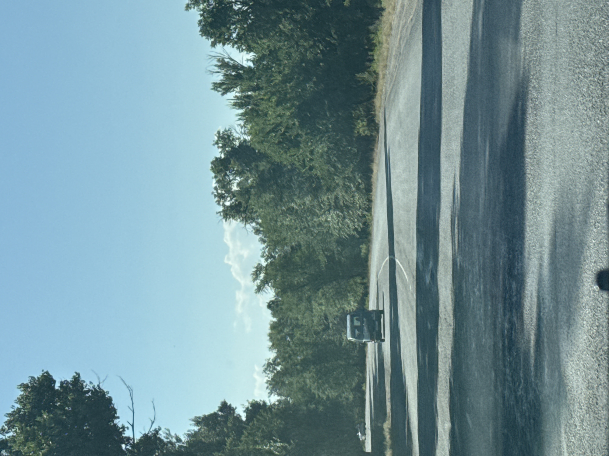 The image shows a paved road with a van driving on it. The road is bordered by trees on both sides, and there is a blue sky with some clouds above. The scene appears to be in a rural or suburban area. There is no indication that this image is from the city of Aral, Kyrgyzstan.