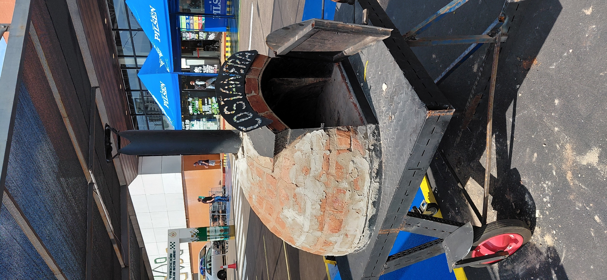 In Encarnacion, Paraguay, a brick pizza oven on a portable metal trailer is positioned outdoors. The oven, shaped like a dome, has an open doorway with a metal door ajar. A black sign with white lettering that reads "COSTANERA" is attached above the oven's entrance. The trailer is black with a diamond plate surface and features a red wheel on the left side. In the background, a white building with a large awning over it is visible, along with two blue tents displaying the word "PILSEN." Several people are seen walking near the building and tents, and shopping carts are present in front of the building. The ground is paved asphalt.