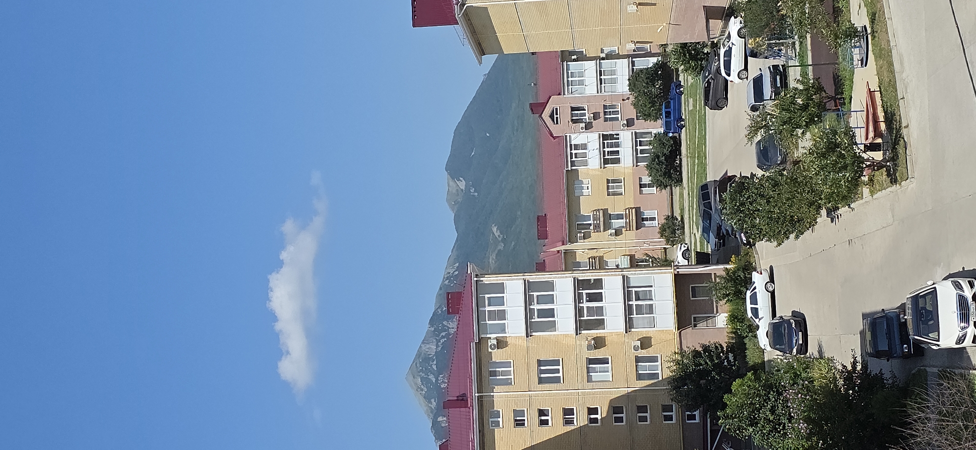 The image depicts a residential area in Inozemtsevo, Russia, with multi-story apartment buildings in the foreground and a mountain range in the background under a clear blue sky with a single cloud. The apartment buildings are light yellow with red tiled roofs and white window frames and balconies. Several cars are parked in the paved area between the buildings, including a white sedan, a dark sedan, a black SUV, a blue SUV, and a silver sedan. Green trees and bushes are interspersed around the buildings and parking area. A small playground with a sandbox and a merry-go-round is visible to the right. The mountains in the background are covered in green vegetation, with some exposed rocky areas and patches of snow near the peaks. The overall scene is a daytime view of a suburban environment.