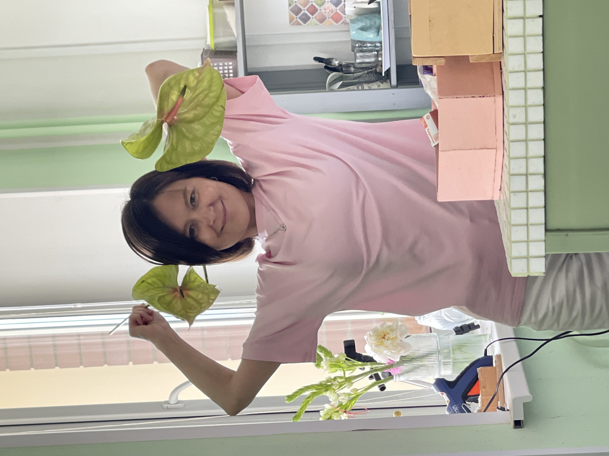 A woman in a pink t-shirt is standing in front of a window, holding up two light green anthurium flowers. She is smiling at the camera, her head tilted slightly. Her dark hair is short and falls to her chin. Behind her, a shelf unit is visible, filled with various items. On the shelf, there are some decorative tiles, a basket, and what appears to be a glue gun with wires attached. Below the shelf, a vase with white flowers is placed on the window sill. The light appears to be natural, suggesting it might be daytime. The scene is brightly lit, possibly indicating a sunny day. The background is blurred, focusing attention on the woman and the flowers. The overall mood seems cheerful and relaxed.