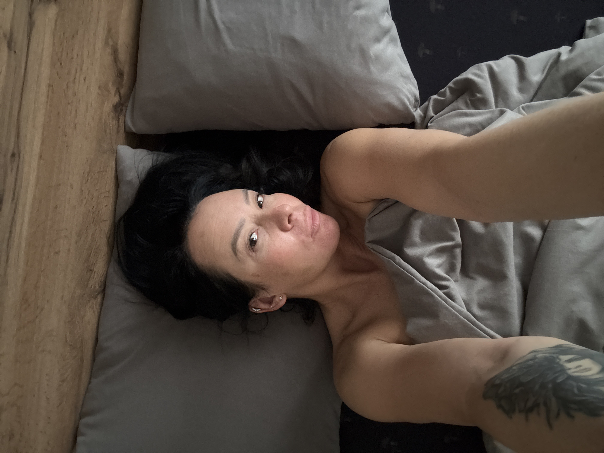 A woman with dark, loosely curled hair is depicted lying in bed, looking directly at the camera with a relaxed expression. Her lips are slightly parted, and she has visible ear piercings in her left ear. Her bare shoulders and upper chest are partially covered by a gray duvet or blanket. A prominent, dark tattoo, possibly featuring wings or a mythical creature, is visible on her left upper arm. Her arm is extended, suggesting she is taking a selfie from an overhead perspective.
The setting is an indoor bedroom. To the left of the woman, a textured wooden headboard or wall paneling runs vertically, providing a warm, natural contrast to the cool tones of the bedding. The bed is furnished with two light gray pillows and a matching gray duvet, which drapes around her. A darker sheet or mattress is visible beneath the pillows. The scene conveys a moment of intimacy and relaxation, likely occurring in the morning or evening. There is no visible text in the image. This scene takes place in Chaikovsky, Russia.
