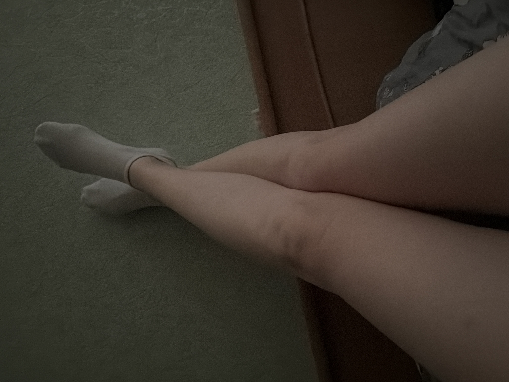 The image shows a pair of legs, one crossed over the other, clad in white socks, resting on what appears to be carpet or a textured surface. A portion of a bed or furniture is visible on the right side of the frame. No further location or city details are discernible from the image.
