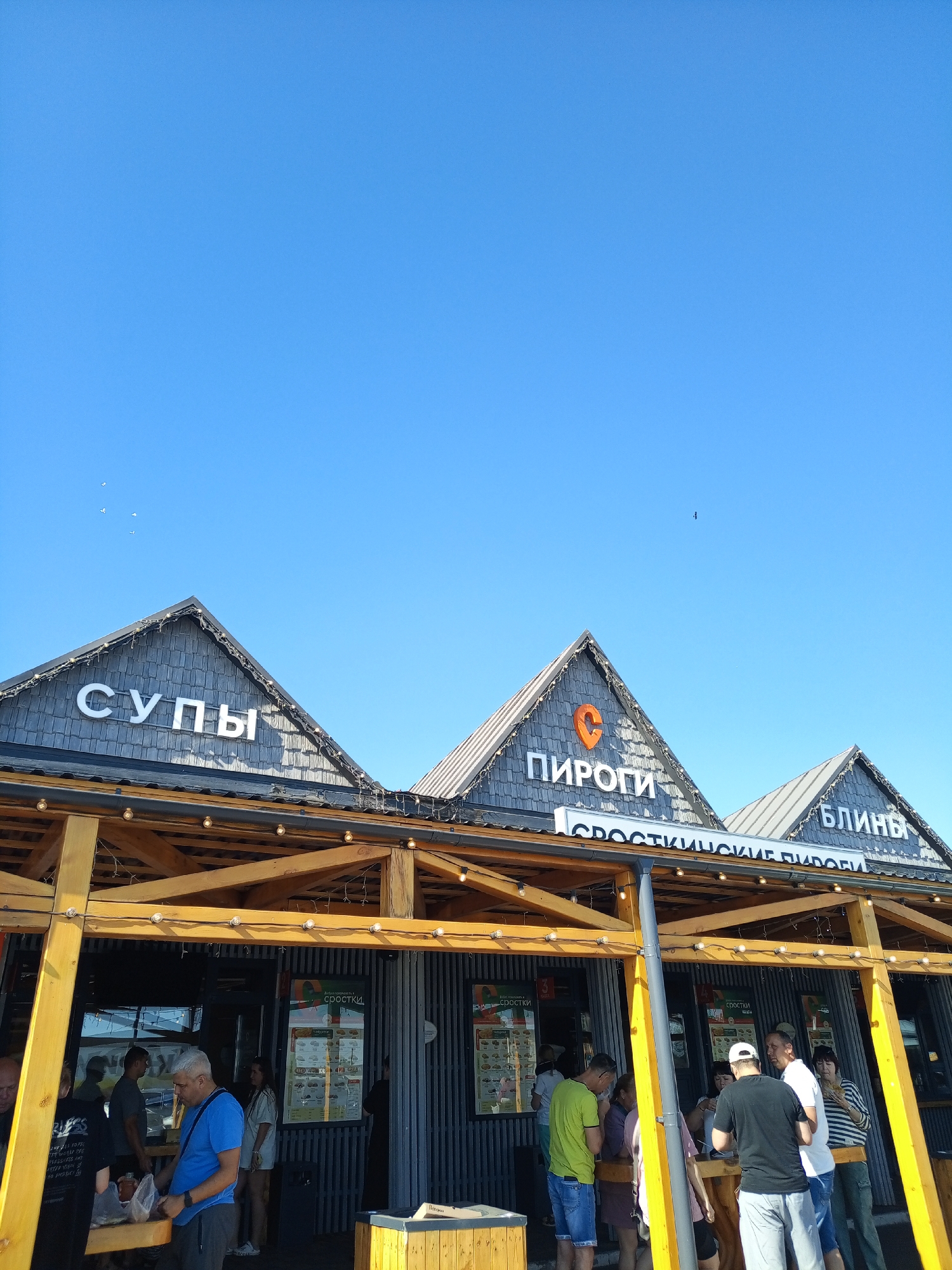 In the city of Srostki, Russia, a bright, clear blue sky dominates the upper portion of the image. Below the sky, a row of buildings with triangular, dark grey roofs is visible, adorned with strings of lights. White, illuminated signs on the roofs read "СУПЫ" (Soups) on the left, "ПИРОГИ" (Pies) in the center with an orange circular logo, and "БЛИНЫ" (Pancakes) on the right. A lower sign spanning across the central and right buildings reads "СРОСТКИНСКИЕ ПИРОГИ" (Srostki Pies).

The foreground features an open-air food establishment constructed with a yellow wooden frame and a dark grey corrugated metal facade. Several people are gathered around counters or standing in small groups. On the left, a man in a blue t-shirt and grey pants is interacting with a table. To his right, a man in a black t-shirt is partially visible. In the center, a group of individuals, including a man in a green t-shirt and denim shorts, and a woman in a light-colored shirt, are engaged in conversation. Further to the right, a group includes a man in a black t-shirt and white baseball cap, a man in a white t-shirt, and a woman in a striped shirt. Men and women in casual attire are observing menus displayed on the walls or interacting with each other. The overall scene depicts a lively outdoor food service area on a sunny day.