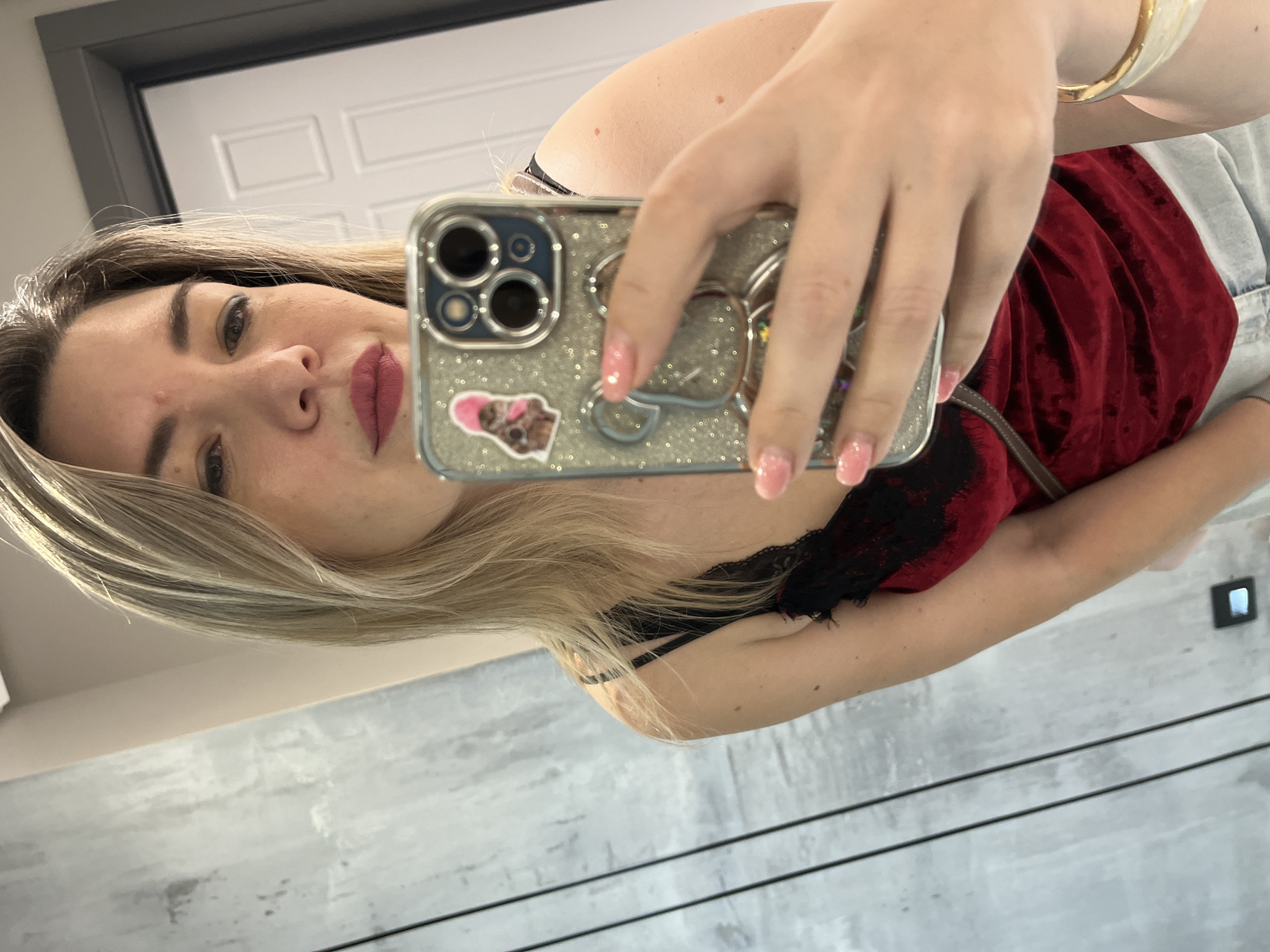 A woman is taking a selfie in a room. She has blonde hair and is wearing a red velvet top with black lace trim and denim shorts. She is holding a silver, glittery phone case with a teddy bear design and a sticker of a cat wearing a pink hat. Her nails are painted pink. The background appears to be a modern interior with a gray, textured wall and a white door. The image was taken in Sochi, Russia.