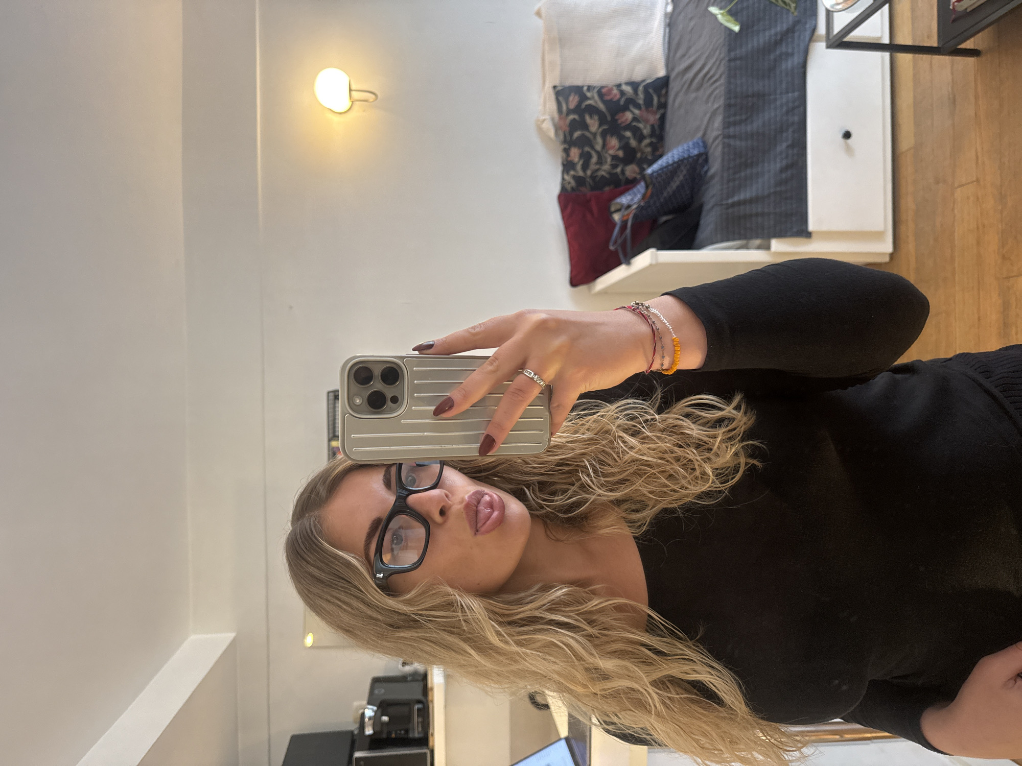 A woman with blonde wavy hair and glasses is taking a selfie. She is wearing a black top and a ring on her finger. She is holding a phone with a gray textured case. In the background, there is a light on the wall, pillows on a shelf, and a wooden floor. There is also some office equipment in the reflection of the mirror she is using to take the photo. The image does not definitively indicate it was taken in Paris, France.