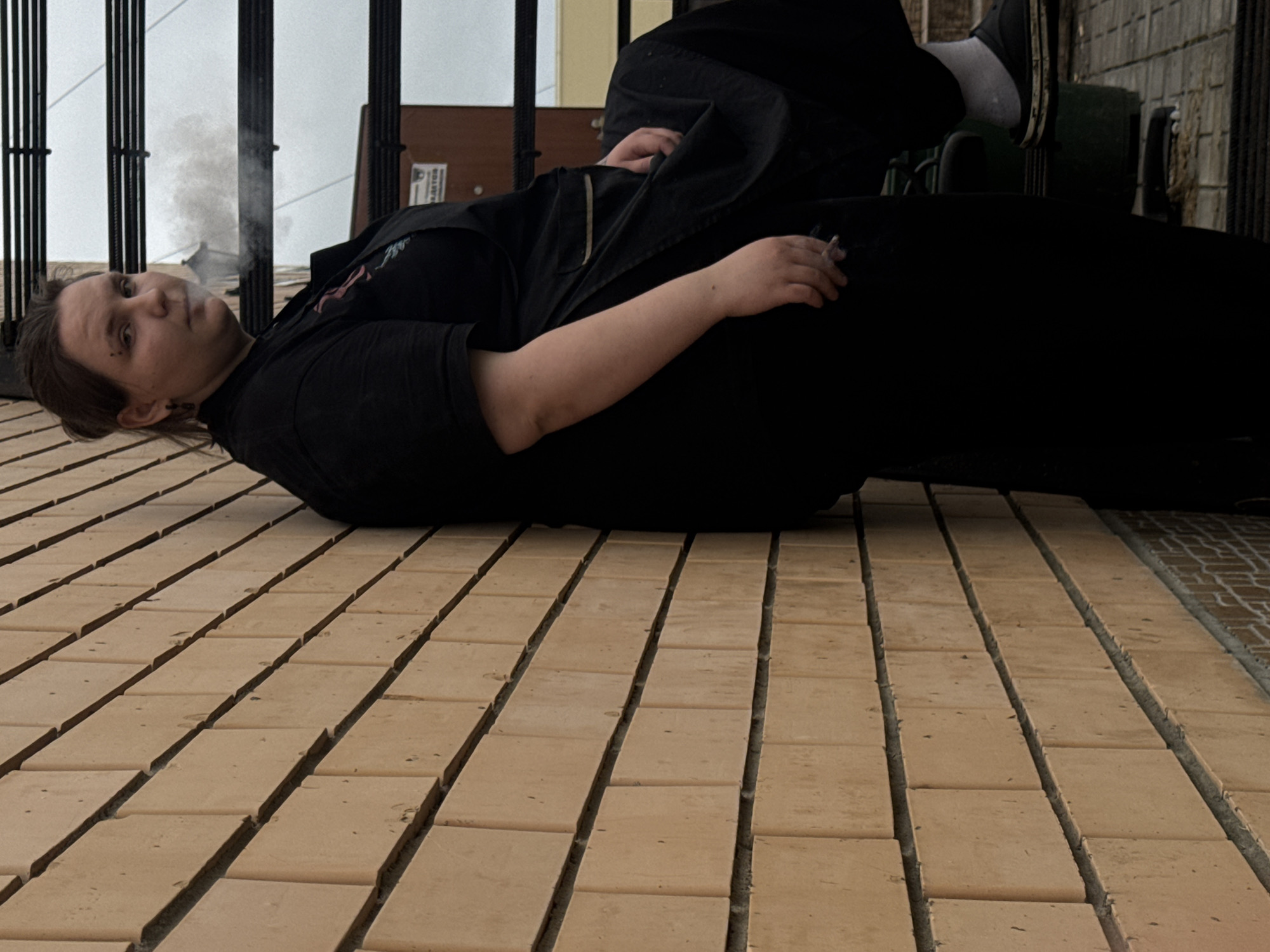 A person is leaning against a light brown brick wall outdoors, exhaling smoke from a cigarette. They are wearing a black t-shirt and black pants, with a black apron over the t-shirt. The person has dark hair pulled back and is wearing earrings. The background includes dark metal grating and some signage, suggesting an urban or industrial area. The ground appears to be paved. The overall impression is a candid moment of someone taking a smoke break against a textured wall.