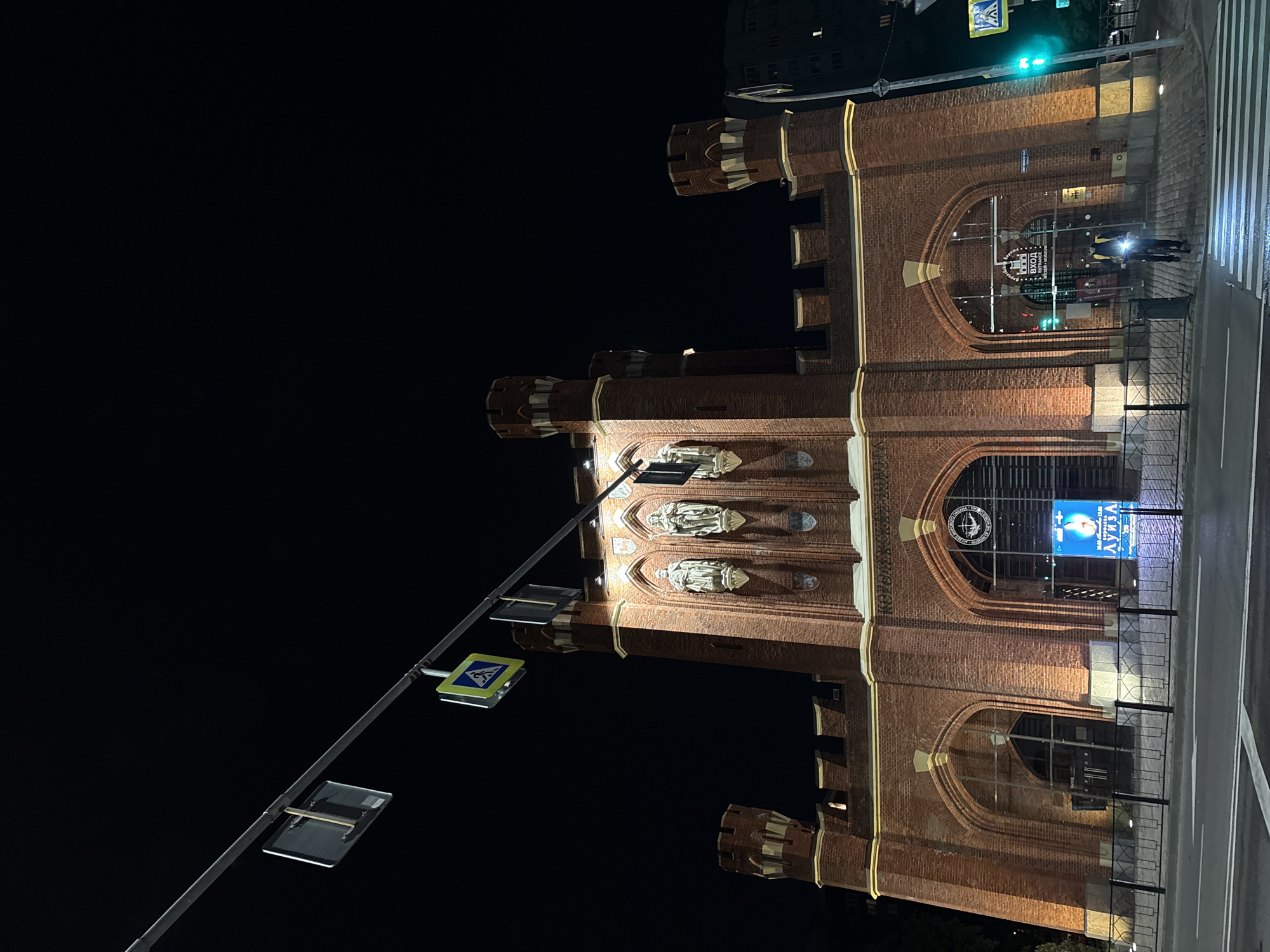 The media file presents an impressive red brick building at night, illuminated against a dark sky. The original image is oriented vertically (rotated 90 degrees clockwise). The structure features multiple crenellated towers and turrets, giving it a fortress-like or historical gatehouse appearance, characteristic of European architecture. Several arched openings are visible, some with large windows or glazed sections.
On the building's facade, there are multiple statues housed within niches, particularly on the upper part of the central section. A street with a marked pedestrian crossing and a sidewalk runs in front of the building, separated by a low metal fence. Street furniture includes a pole with a lit green traffic signal and a yellow square sign indicating a pedestrian crossing.
Visible text on the building includes a circular emblem, likely a logo or crest, and beneath it, clear Russian text "ВХОД" (Entrance) with "ENTRANCE" written below in English, marking an entry point. To the right, a prominent, brightly lit digital display or advertisement board showcases a human face (likely a woman) and the Russian word "ЛУИЗА" (Louisa), along with other smaller, illegible text.
The scene is captured at night, with the building's warm lighting contrasting with the dark surroundings. One person is distinctly visible on the sidewalk near the entrance, seemingly looking towards the building or displays. The architectural style combined with the visible Cyrillic text and the "ENTRANCE" sign suggests the building serves as a public institution, possibly a museum or cultural center, located in Kaliningrad, Russia. The atmosphere appears calm and peaceful under the night sky.