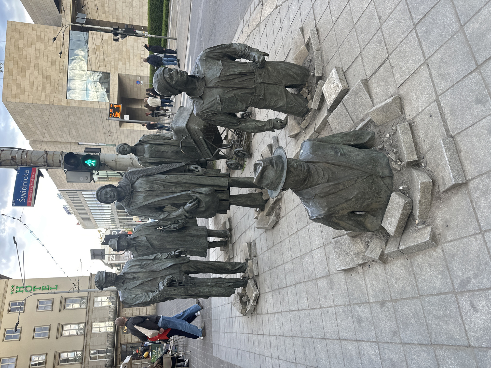 The image shows a bronze sculpture in Wrocław, Poland, known as "Passage" or "Going Through". The artwork depicts a group of figures emerging from or disappearing into the pavement. The sculptures are located at the intersection of Świdnicka Street and Kazimierza Wielkiego Street. The figures are depicted in a realistic style, with some partially submerged in the ground, creating an illusion of them emerging from below or sinking into the pavement.
Several buildings are visible in the background, including a building that is a hotel. People are walking and standing around. A traffic light is visible at the corner.
