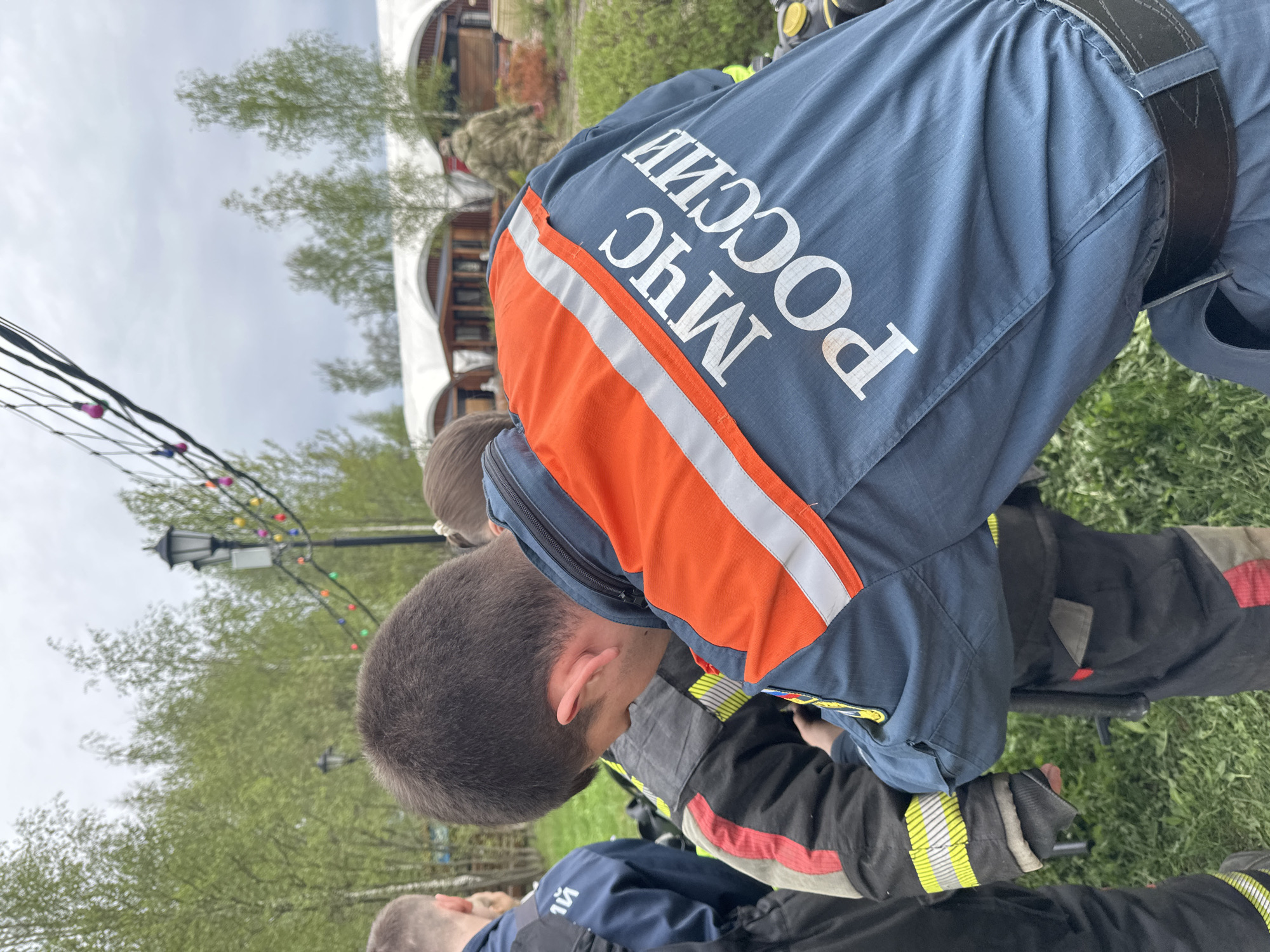 In Grebnevo, Russia, a first responder wearing a dark blue uniform with orange stripes and the Russian acronym for Ministry of Emergency Situations (МЧС РОССИИ) is bending over a person in a firefighter's turnout coat. The two are located on a grassy area near a lamppost with strings of lights. A building with white curved supports is visible in the background.
