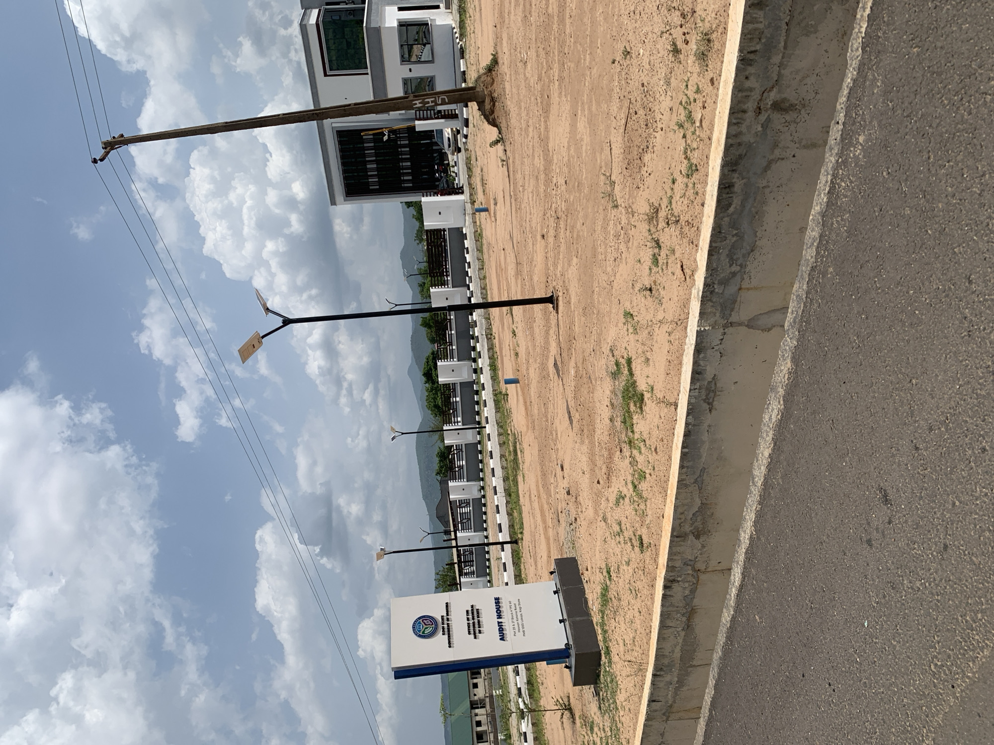 The image depicts an outdoor scene in Lokoja, Nigeria. A sign for "Audit House" is visible, with the address: Plot 25 & 27 Block 4 TPS 60, Katsina Road, PMB 1055 Lokoja, Kogi State. In the background are a modern building, street lamps, and trees. There is a dirt area adjacent to the road. Overhead, there are power lines and a partly cloudy sky.