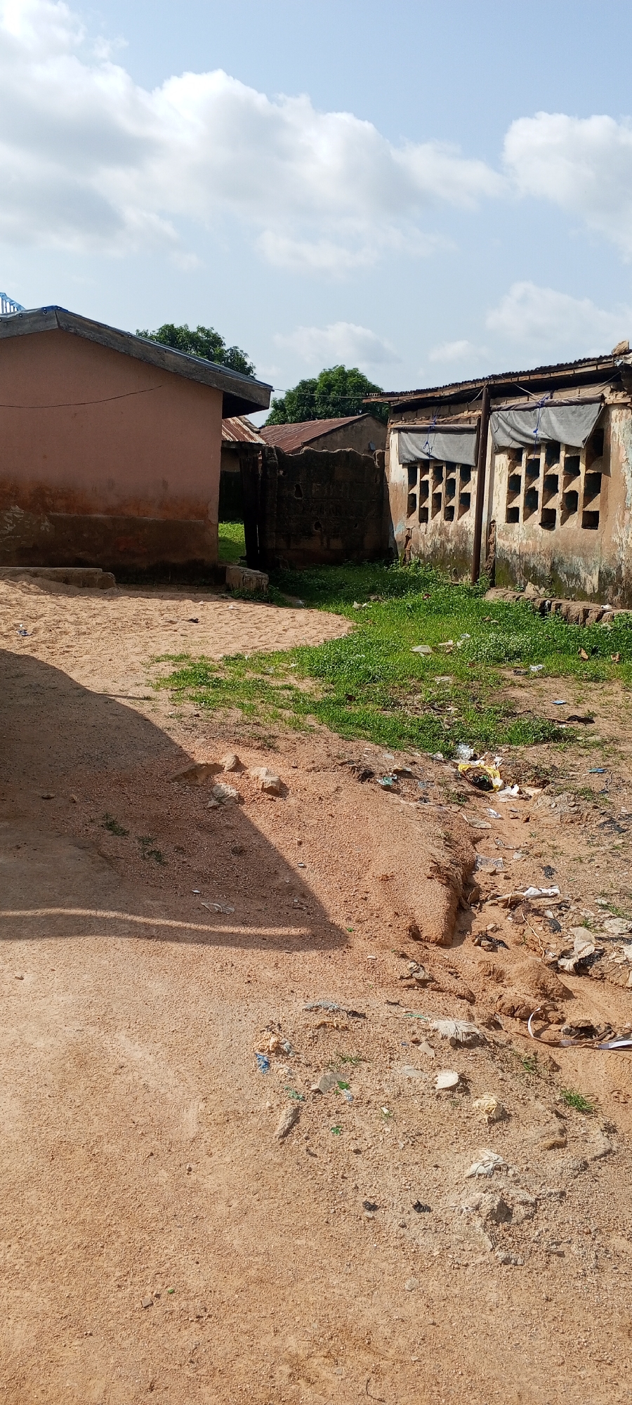 The image depicts an outdoor scene in Jalingo, Nigeria, focusing on a weathered building with a dusty, sandy foreground. The building, likely a residential or commercial structure, exhibits signs of age with peeling paint and a dilapidated roof. Its walls are a faded pinkish-brown, and there are several openings in the right side of the building that resemble ventilation shafts or windows. The foreground is primarily composed of reddish-brown sand, with patches of dry grass and scattered debris, including plastic bags and paper waste, indicating an unkempt environment. A dark shadow falls across the sandy area from an unseen object. In the background, more structures with corrugated metal roofs are visible, along with some green foliage from trees. The sky above is partly cloudy, with white clouds against a pale blue backdrop. The overall impression is one of a humble, possibly rural or semi-urban, setting in Nigeria.
