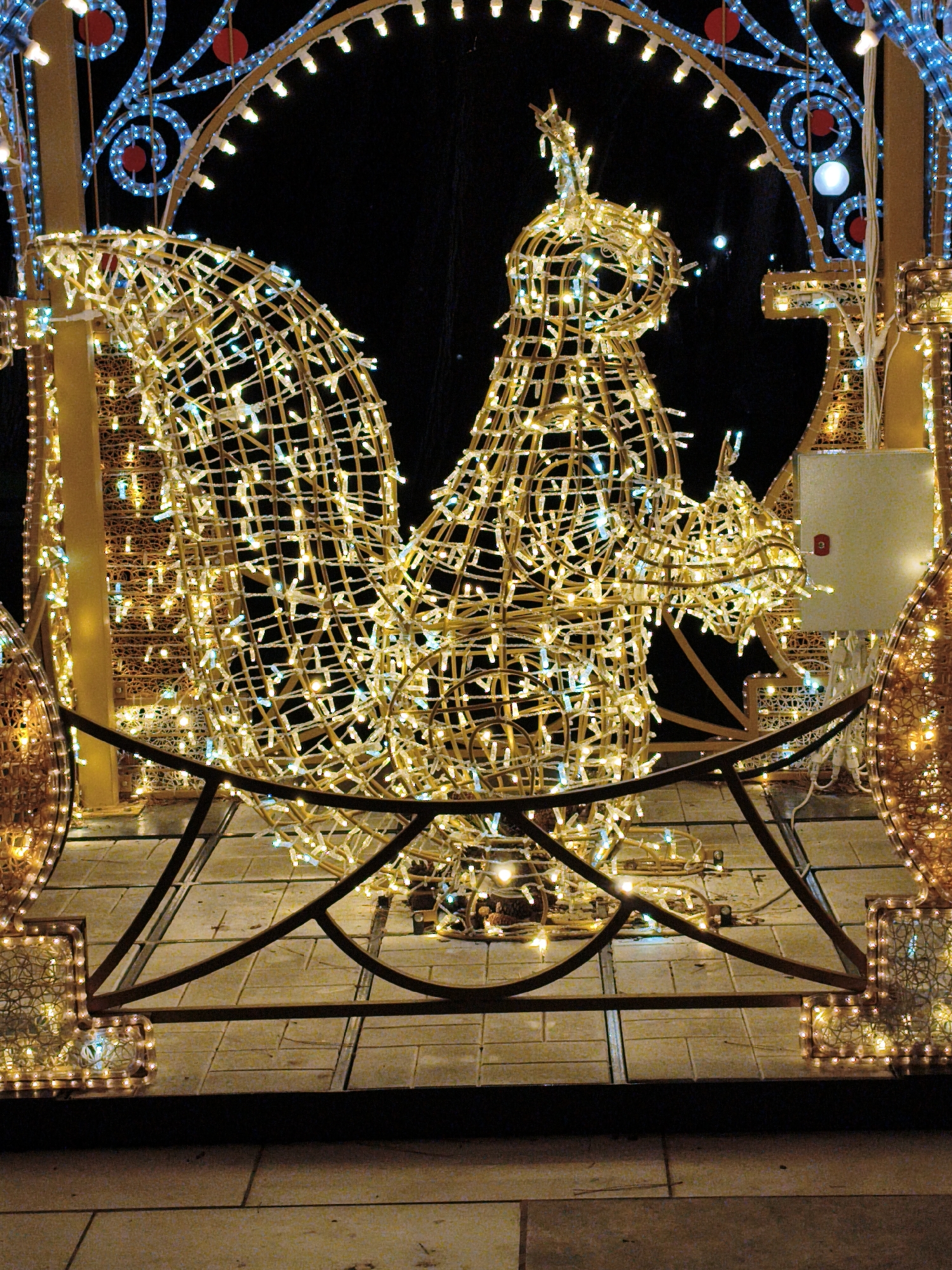 The image depicts an outdoor night scene in Divnomorskoe, Russia, featuring elaborate Christmas or New Year's illuminations. The central and dominant subject is a large, wireframe sculpture of a squirrel, intricately wrapped in strings of warm white and occasional blue lights, creating a glowing effect against the dark background. The squirrel sculpture is positioned behind a dark, decorative metal barrier. To the left and right of the squirrel are additional illuminated structures, including what appears to be a portion of a large archway adorned with blue lights and red spherical accents. The ground is paved with light-colored tiles. The overall impression is one of festive holiday decoration in a public space. There are no people or animals visible in the frame.