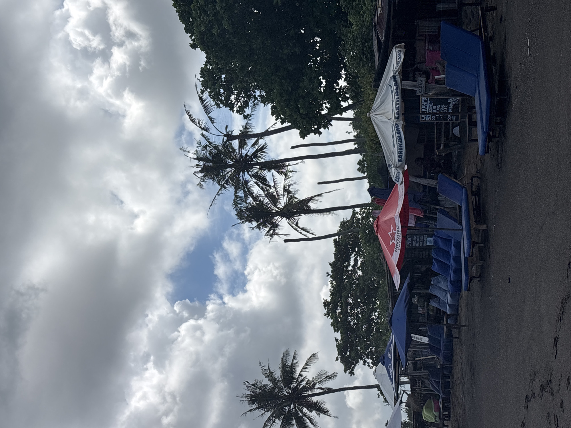 This is an outdoor shot taken at a beachside location in Mengwi, Indonesia. The viewpoint is from ground level looking upwards towards the sky, capturing a series of tall palm trees with their fronds swaying in the breeze. The sky is filled with a mix of puffy white and dark gray clouds, with patches of blue peeking through.
Along the right edge of the image, there is a row of beach umbrellas, some closed and some partially open, providing shade. These umbrellas feature various colors, including white, red, and blue. Several lounge chairs are situated beneath the umbrellas, suggesting a place for relaxation and enjoyment of the beach.
In the backdrop, there are also other trees with denser, greener foliage. This area appears to be a spot with restaurants, vendors or bars on the beach. The photo composition also captures signs and structures that give context to this area.