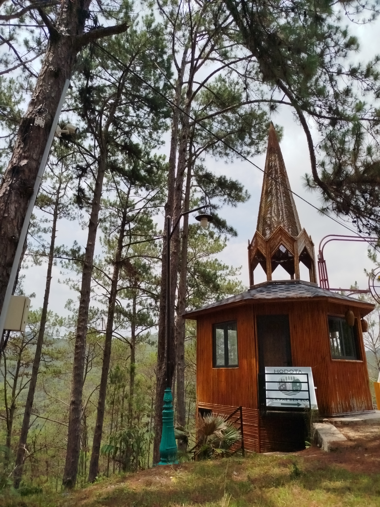 This eye-level outdoor shot features a unique wooden building with a prominent spire, situated amidst a dense pine forest in Păng Tiêng 1, Vietnam. The building, constructed from horizontal wooden or bamboo panels, has dark-framed windows and a shingled roof, topped by an intricately woven, multi-tiered spire that resembles a traditional pagoda.
A white sign, attached to the building's front, displays the name "HODOTA" at the top. Below this, a logo depicts pine trees, water, and mountains, followed by the Vietnamese text "CÀ PHÊ BÊN SÔNG" (Coffee by the river) and an arrow pointing right, indicating a coffee shop or similar establishment. To the left of the building, a decorative green post or base of a lamp is visible on the grassy ground.
The scene is dominated by numerous tall, slender pine trees with green needles, their trunks rising high into an overcast or partly cloudy sky. A security camera is mounted on the trunk of a large pine tree on the far left, accompanied by wires and a set of keys hanging nearby. Another white utility box is attached lower on the same tree. A black lamp post with a light fixture stands to the left of the building, extending above its roofline. The background reveals a hilly landscape covered in more trees, suggesting a mountainous or forested region.
The image portrays a peaceful, natural setting under diffused daylight, with no people visible. The primary colors are the greens of the trees, the browns of the building and tree trunks, and the grey of the sky.