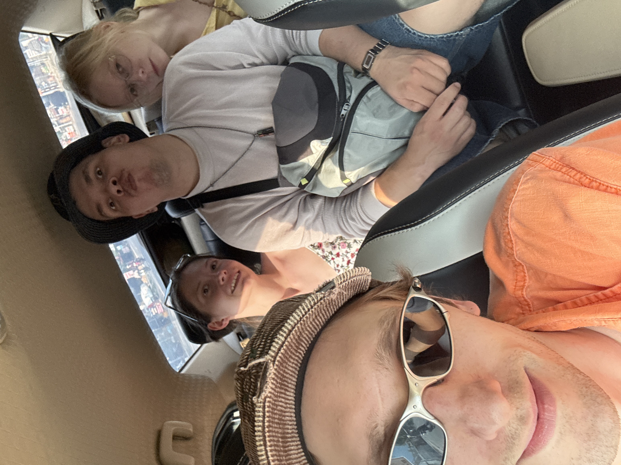 Four individuals are positioned within the confines of a vehicle's interior, likely in motion through the city of Tay Nha Trang Ward, Vietnam, as evidenced by the visible urban landscape through the rear window. The foreground is dominated by the face of an individual wearing silver-framed sunglasses and an orange collared shirt. Behind them, to their left, sits another person wearing a patterned cap and a floral-print top, with a faint smile. In the middle row, a person sporting a dark bucket hat makes a puckered facial expression, their torso adorned with a grey and black crossbody bag and a necklace. To their right, a blonde individual wearing glasses and a yellow top leans their head towards the other person. The atmosphere appears casual and informal, consistent with a group traveling together.