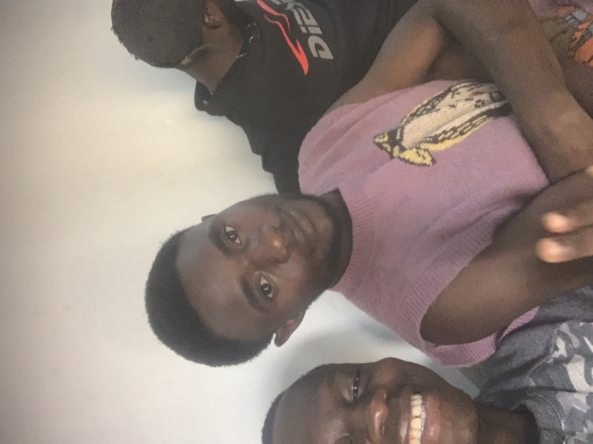 Three young, Black people are positioned in close proximity, suggesting a selfie. The individual closest to the camera is smiling and wearing a gray patterned shirt. The person in the middle is wearing a mauve sweater with a decorative pattern. The third person is wearing a black hoodie with the word "Diesel" on the front. The background is a plain white surface, likely a wall. The location is possibly Gura Top, Nigeria.
