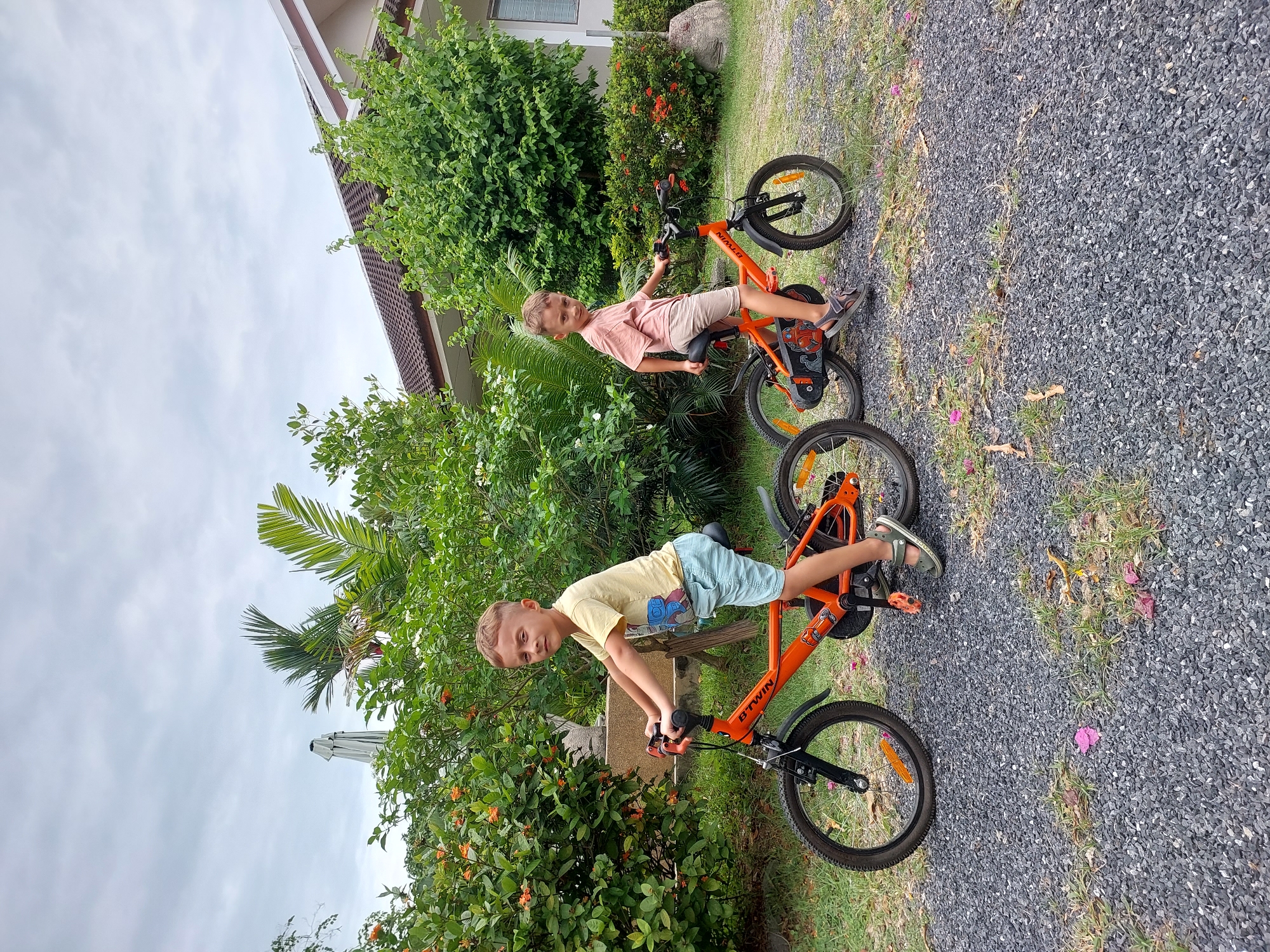 Two young boys are sitting on orange bicycles in an outdoor setting. The boy on the left is wearing a yellow t-shirt and light blue shorts, while the boy on the right is wearing a pink t-shirt and beige shorts. They are positioned on a gravel path in front of lush green bushes and a building with a tiled roof. The sky is overcast. The location is the Ko Samui District in Thailand.