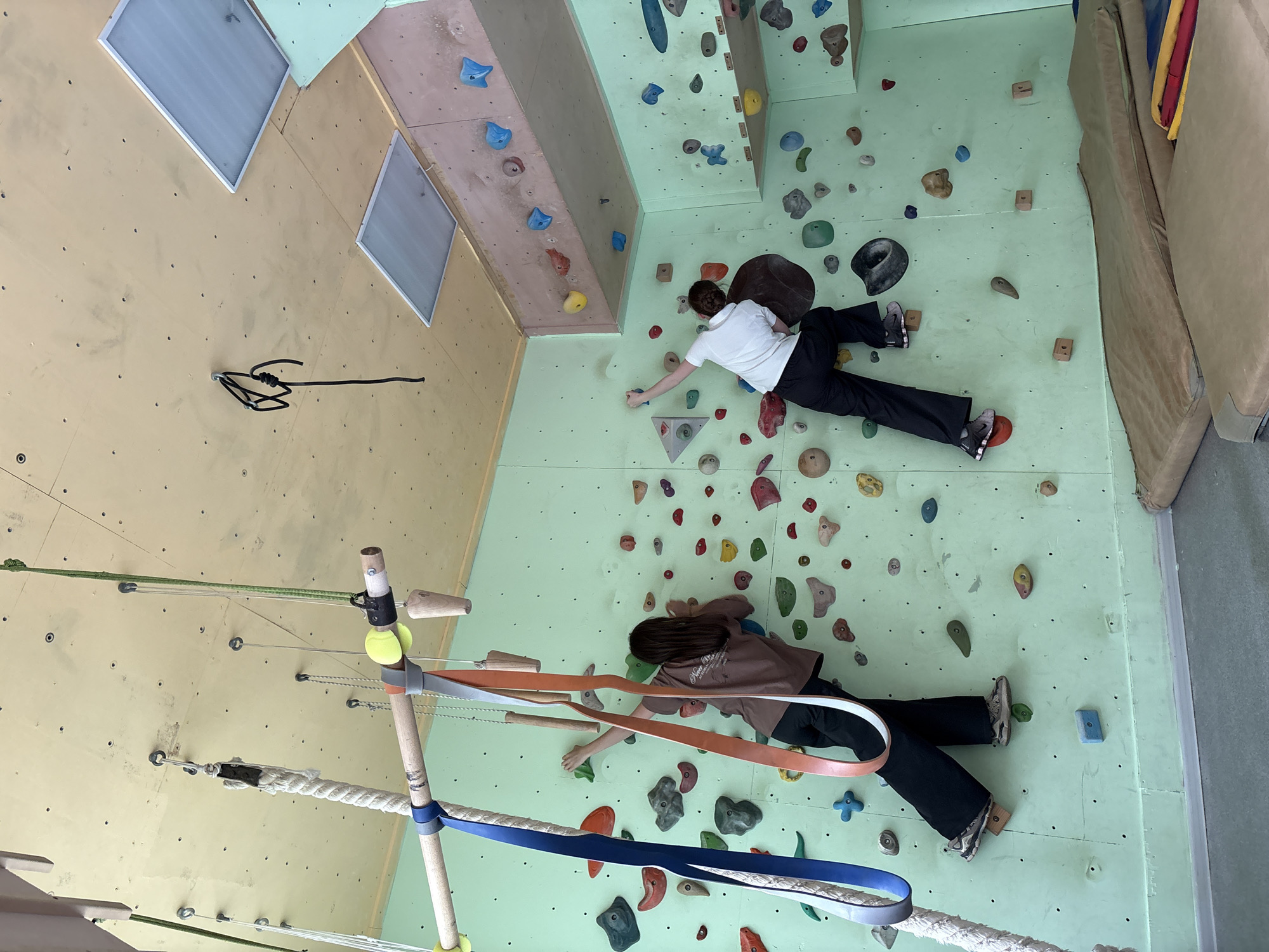 Two people are participating in what appears to be a modified climbing exercise within an indoor climbing gym. One person is wearing a white shirt and black pants. The other person is wearing a brown shirt and black pants. Both individuals are positioned horizontally on a light green climbing wall. The wall features various colored climbing holds. A wooden structure with ropes and straps is in the foreground. The wall to the left is yellowish. There are three light fixtures in the left corner of the room. Based on the prompt, this could be located in Chaikovsky, Russia.