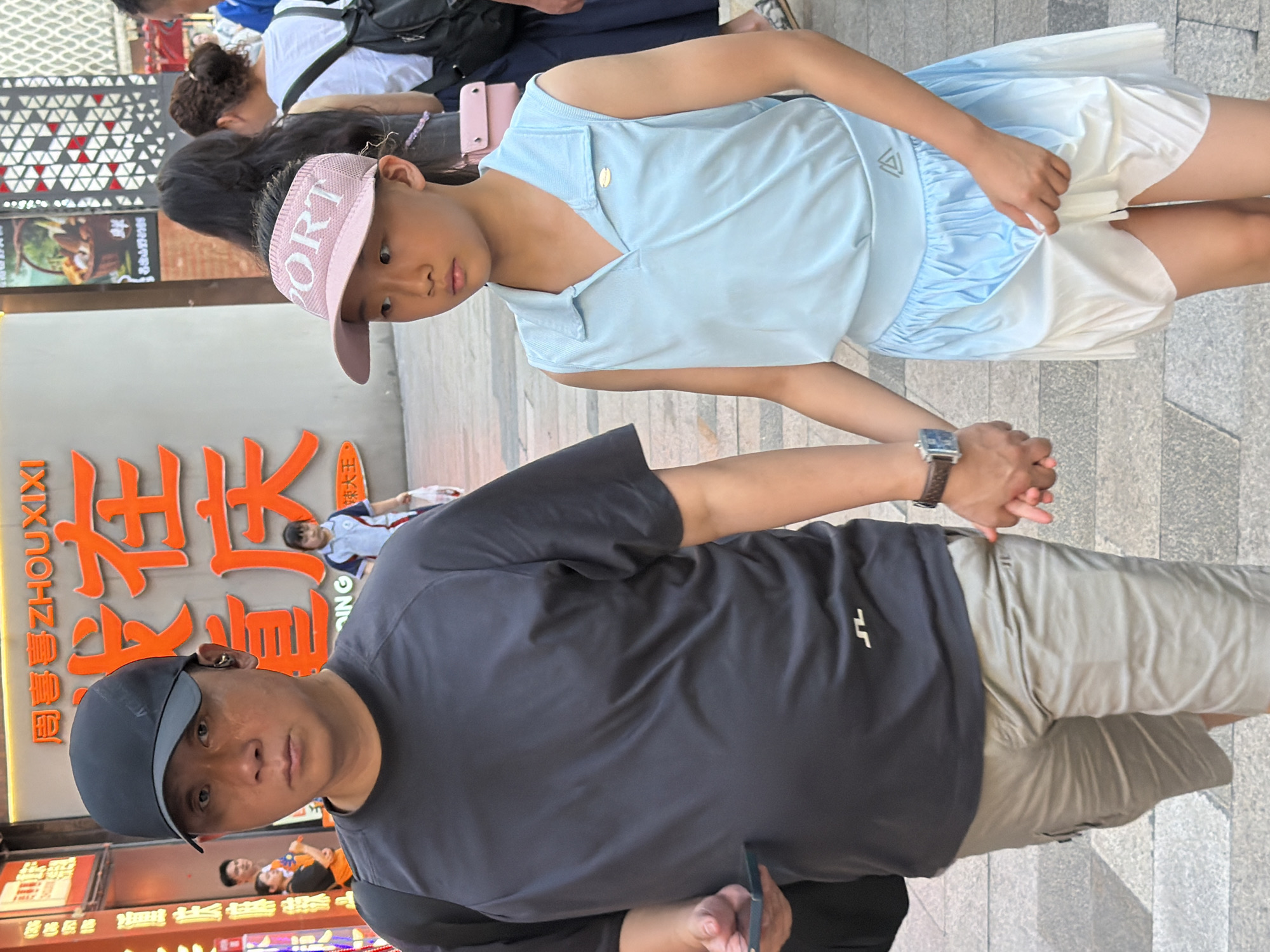 The image shows a vertical shot of several people in an urban outdoor setting. The foreground is dominated by a man wearing a dark grey t-shirt, a dark baseball cap, and light grey shorts. He is looking directly at the camera with a neutral expression. His right hand is holding a smartphone. To his left, a young girl is visible, wearing a light blue sleeveless top and a white pleated skirt. She has a pink visor with the word "SPORT" on it. She is holding the man's hand. In the background, a large orange sign with Chinese characters is prominently displayed. More people are visible in the background, including a woman with dark curly hair and others partially obscured. The setting appears to be a street or plaza, with a paved ground and a patterned wall in the upper left corner. The lighting suggests it is daytime. The presence of the large sign with Chinese text and the architectural style hint at a location in China, specifically Yuzhong District.
