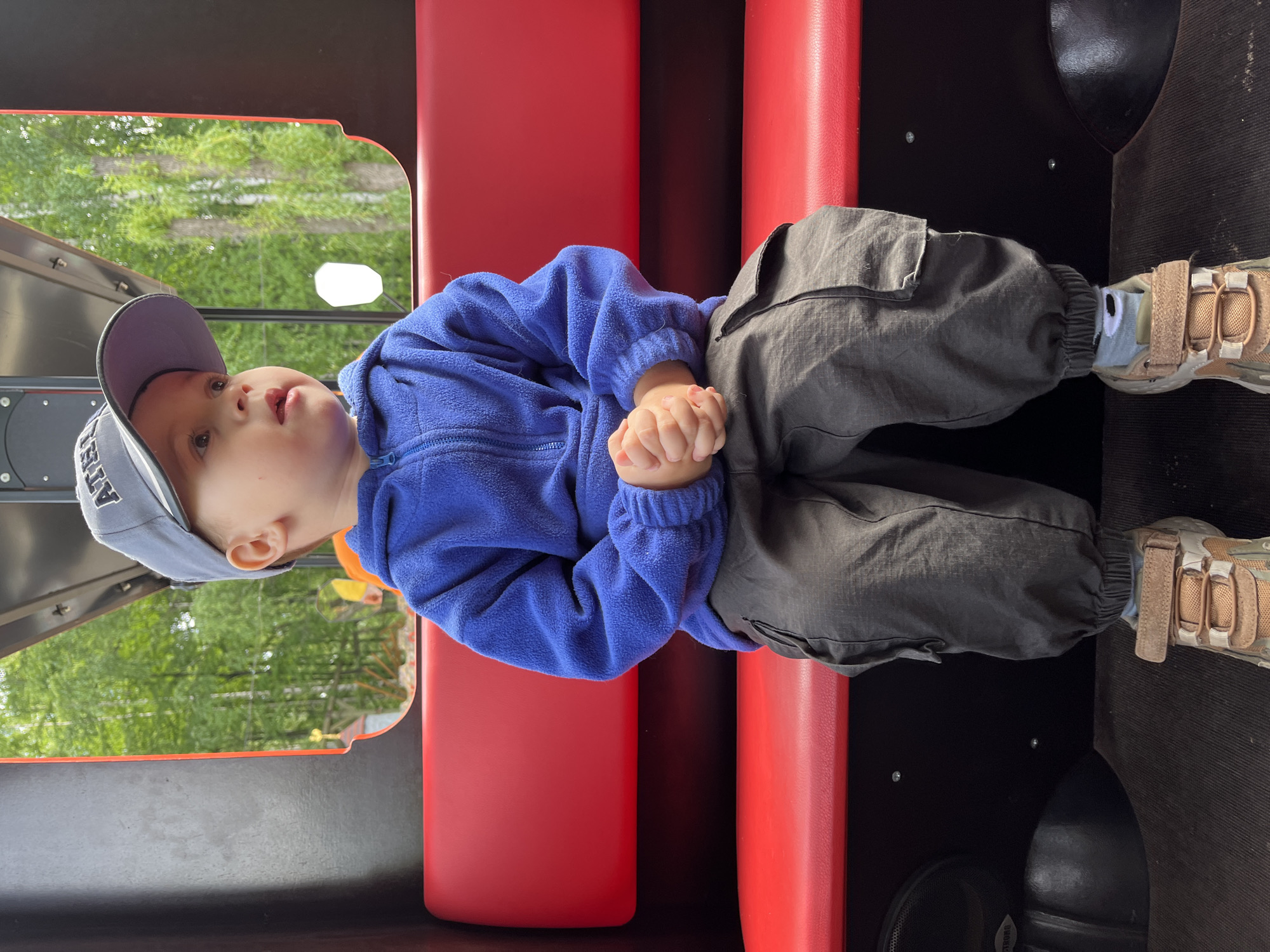 A young child, approximately a toddler or preschooler, is depicted seated inside a train car, likely an amusement park ride, in the city of Chaikovsky, Russia. The child is positioned centrally on a red upholstered bench, with a matching red backrest behind. The interior walls of the car are black, and a dark, textured floor is visible. On the lower left wall, a speaker grill with the text "STRONG" is partially visible.
The child wears a grey baseball cap with dark lettering (partially obscured, showing "AT"), a royal blue fleece full-zip jacket, dark grey cargo pants with side pockets and elasticized cuffs, and light brown and beige athletic shoes with Velcro straps. A light green sock with a white, stylized cartoon face is visible on the left foot. The child's hands are clasped together in front of the lap. The child's head is turned to the right, looking out through a window opening on the right side of the frame.
Through the window, a lush, green environment is visible, dominated by trees, some with light-colored bark. In the background, hints of other structures can be discerned, including a yellow object and a white, hexagonal light fixture, suggesting a recreational or park setting.