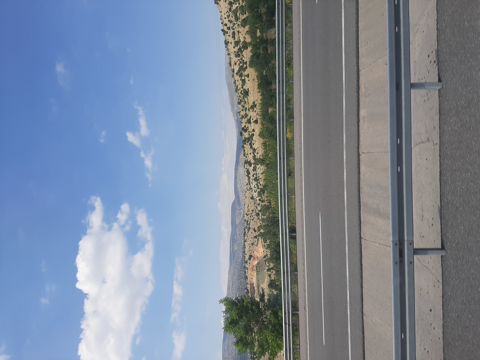 The image captures a view from a moving vehicle on a highway, likely in or near Meram, Turkey, based on the provided context. The scene is dominated by a clear blue sky with scattered white clouds. In the foreground, a multi-lane asphalt road is visible, marked with white lines. A grey metal guardrail runs along the side of the road, separating it from the natural landscape.

Beyond the road, the environment consists of rolling hills covered in dry grass and scattered green trees and bushes. In the mid-ground, a section of the hills appears to be a cleared area, possibly a quarry or a construction site, with exposed orange earth. The background shows a vast, undulating landscape extending towards the horizon. The overall impression is one of a rural or semi-rural setting viewed from a highway on a sunny day. No people or specific actions are depicted in the image.