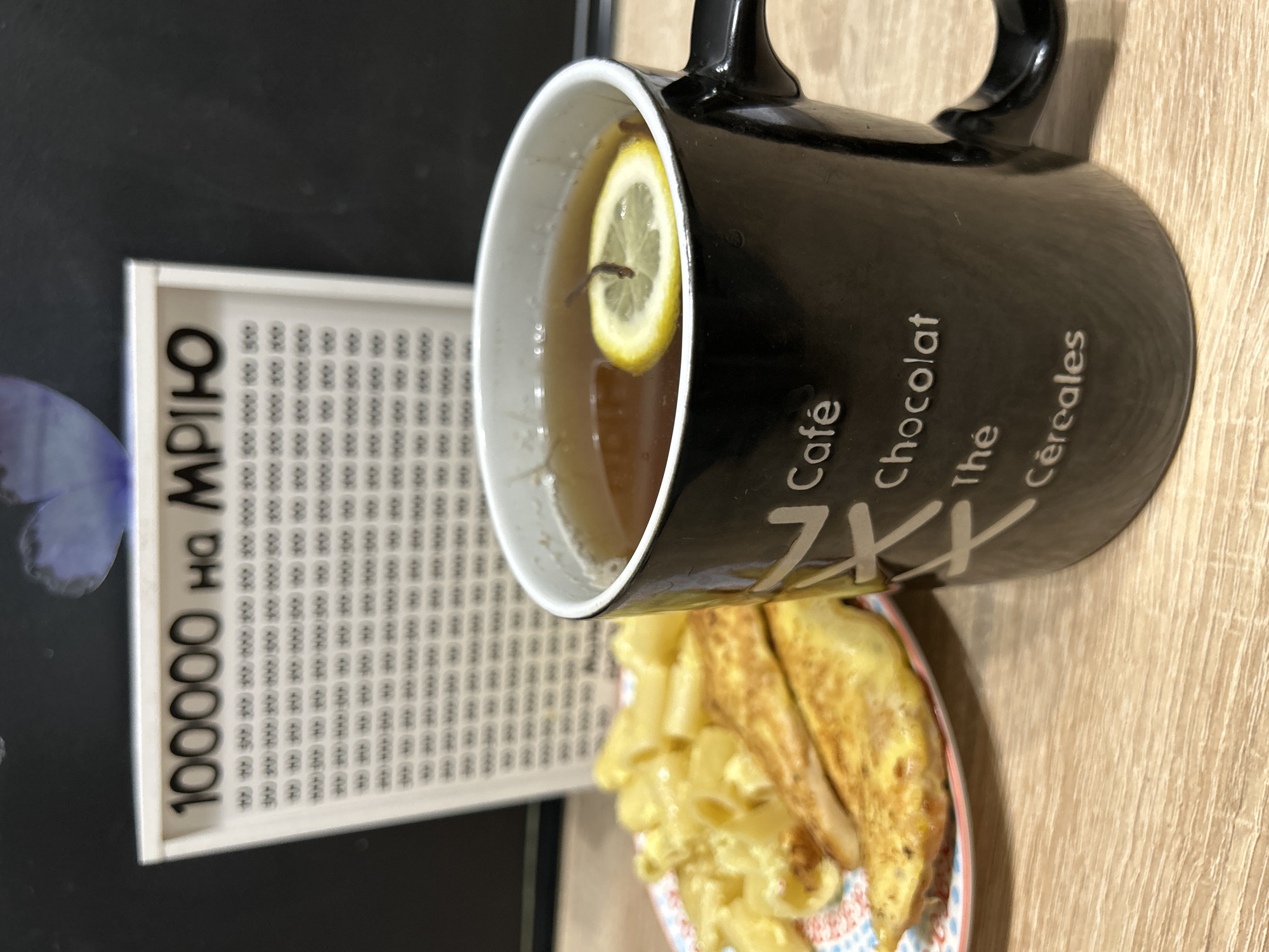 The image shows a close-up of a mug filled with tea, a slice of lemon floating on top, and a plate of food containing pasta and chicken breasts. The mug is black with white text that appears to be in French, reading "Café Chocolat Thé Céréales." In the background, a white board with the text "100000 на мрію" (100,000 for a dream) and a grid of checkboxes is visible. The scene appears to be set indoors on a wooden surface. The location is not specified as being in Ternopil, Ukraine.