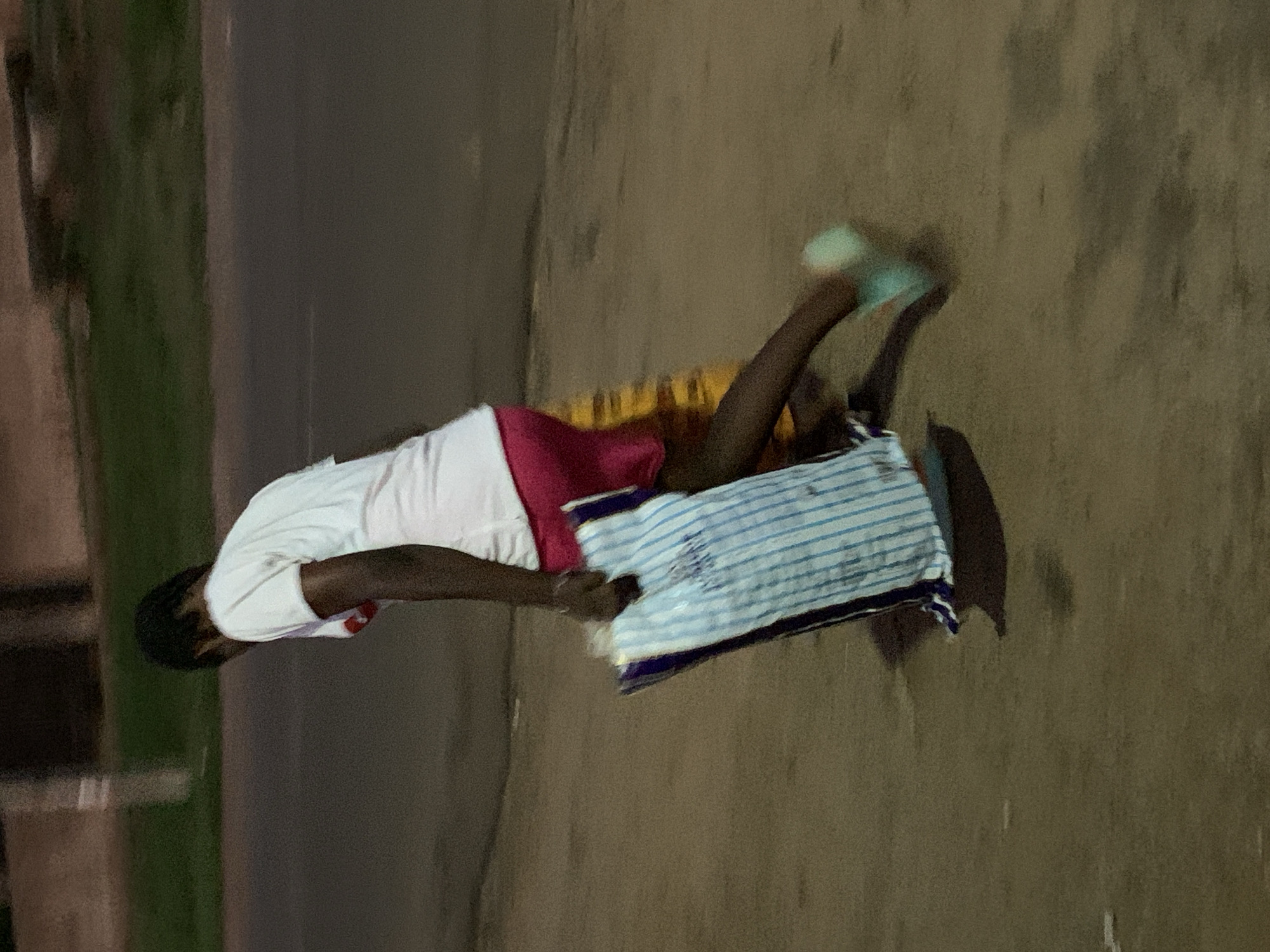 A person wearing a white shirt and pink shorts is lying on a striped pillow on a concrete surface. The person is wearing what appears to be light green footwear. The location appears to be outdoors, possibly in an urban area, but the specific location in Gbeleko, Nigeria, cannot be determined from the image alone. The environment includes a dark asphalt surface and some blurred greenery in the background. The person's actions are limited to lying on the pillow. There are no apparent interactions with other people or objects.