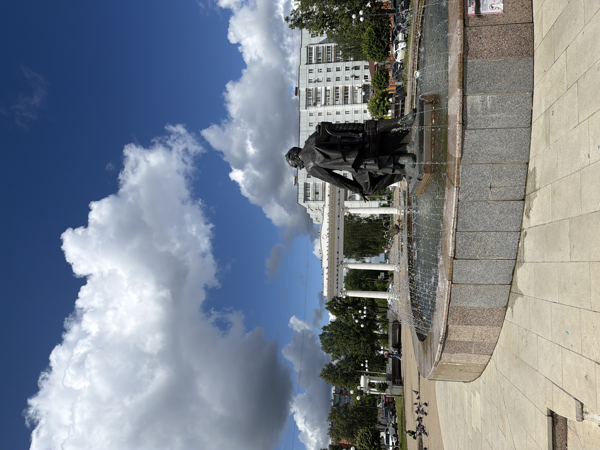 The image shows a statue of Pyotr Ilyich Tchaikovsky located in Chaikovsky, Russia. The bronze statue is depicted seated and is positioned within a fountain structure. Water jets are active within the fountain. Behind the statue, there is a white colonnade structure with multiple columns. A multistory building with a repetitive window pattern is visible in the background. The sky is blue with a mixture of white cumulus and darker cumulonimbus clouds. The foreground shows a paved area with a curved stone edge. A sign with Cyrillic text is attached to the fountain. Pigeons are visible on the ground near the fountain's edge.