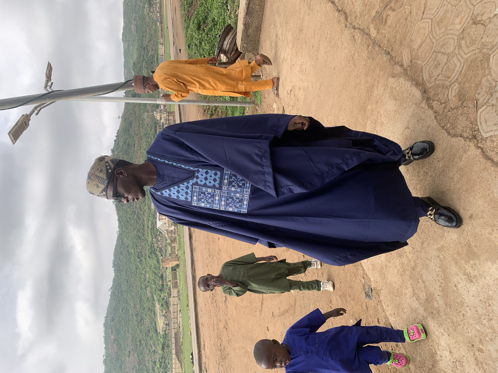 The image shows a man in a dark blue traditional Hausa robe, called a Babban Riga, standing on a concrete walkway in Jos, Nigeria. He is wearing a patterned cap and dark shoes with decorative embellishments. Behind him and to his right are three children, one in a yellow robe, one in a green robe and the other in a dark blue robe. In the background, a hillside with green vegetation is visible under an overcast sky. A light pole is visible to the left of the man in the dark blue robe. In the background, near the hillside, is what seems to be a building site. The ground appears to be a red-brown earth.