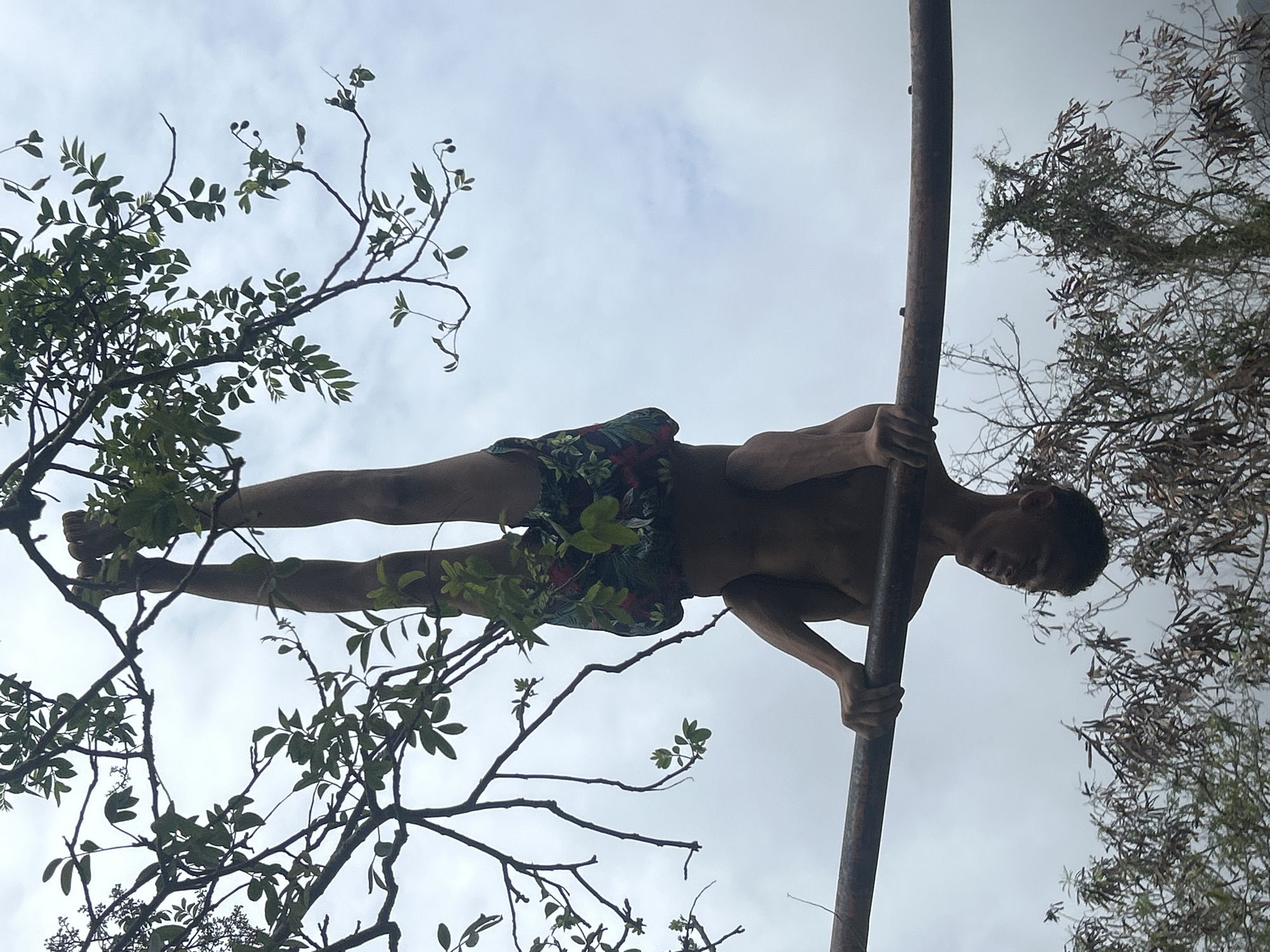 In Pedro González, Venezuela, a single individual, likely male, with a medium-dark complexion, is suspended upside down in an outdoor environment. The person is shirtless and wears patterned shorts featuring green foliage and red accents on a dark background. Both hands grip a horizontal, cylindrical metal bar, which appears rusted and dark-toned, positioned across the lower mid-section of the image. The individual's legs are bent, with the calves and feet hooked around a thick, leafy tree branch positioned above the metal bar, contributing to the inverted posture. The background consists of an overcast, light grey sky with uniform cloud cover. Dense green tree foliage is prominent in the upper frame, partially obscuring the individual's upper body and legs. Below the metal bar, additional trees or shrubs with darker, possibly drier foliage are visible against the sky.