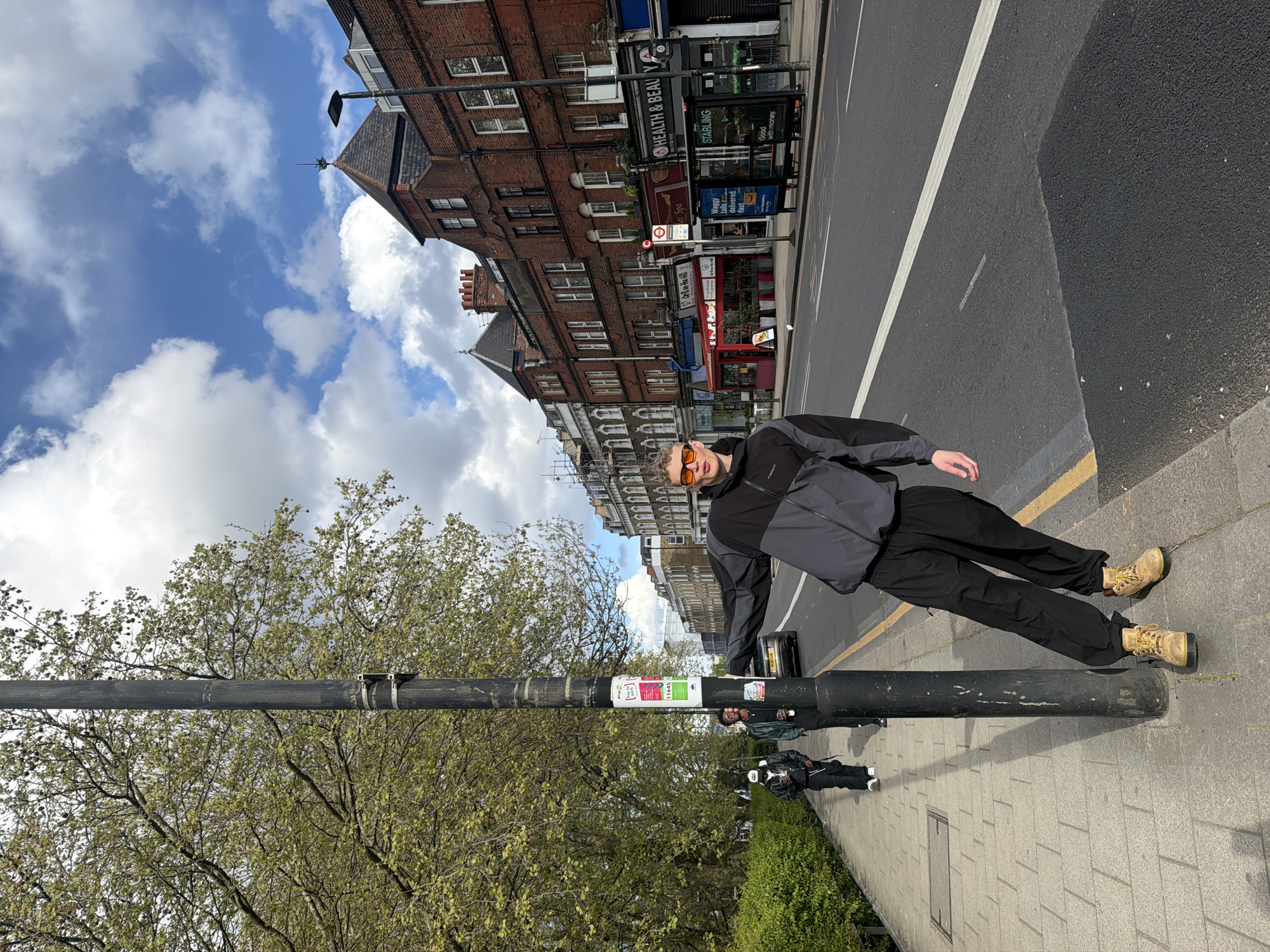 The image depicts a man wearing sunglasses, a two-tone gray and black jacket, black pants, and tan work boots leaning against a black lamppost on a sidewalk. Behind the man is a road and buildings lining the street, with a mix of brick and light-colored facades. The street includes a bus stop and storefronts, with signage for "HEALTH & BEAUTY" visible. There's a partial view of the sky with clouds above the buildings. In the lower left corner, there's a tree with budding leaves, and along the edge of the sidewalk, a narrow strip of greenery is seen. Additional people can be seen further down the sidewalk, and the lamppost has several stickers attached to it. The scene is in London, United Kingdom.