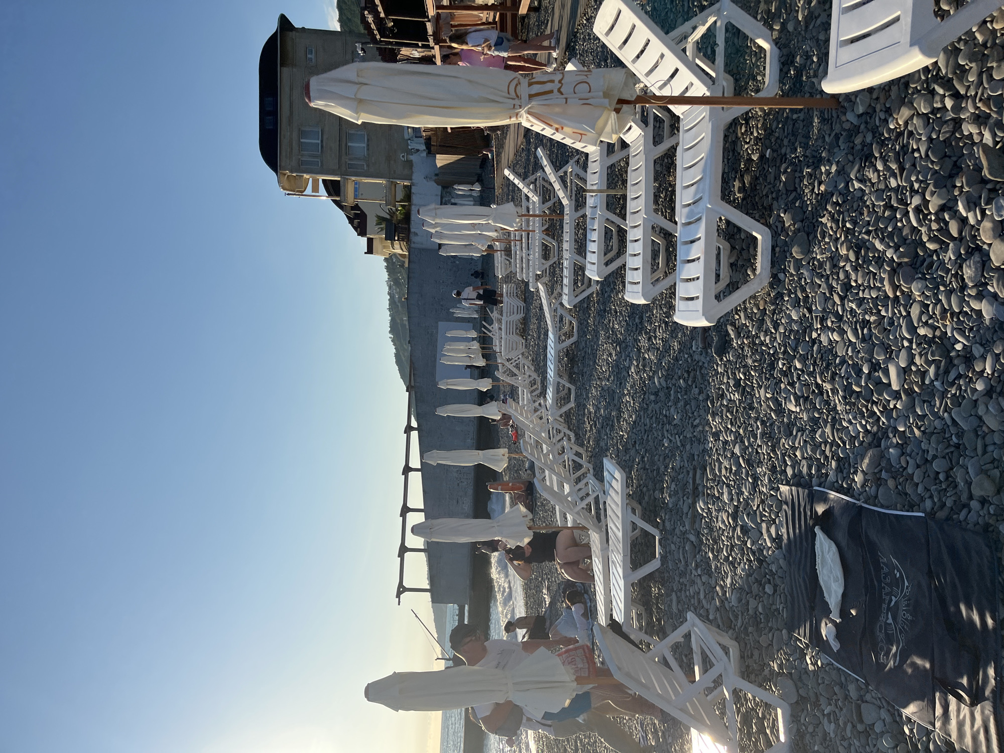 The image depicts a pebble beach in Sochi, Russia, lined with rows of empty white plastic sun loungers and folded white beach umbrellas. In the background, a multi-story building with a dark, curved roof stands against a clear blue sky. Some people are visible on the beach, near the water's edge, where waves are breaking against a concrete embankment. One person is sitting on a sun lounger in the mid-ground, holding their head. On the left side, a man is standing near the water, and further left, a black beach mat with a white towel is laid out on the pebbles. The overall scene suggests a beach resort in the late afternoon or early evening, with the sun casting long shadows.