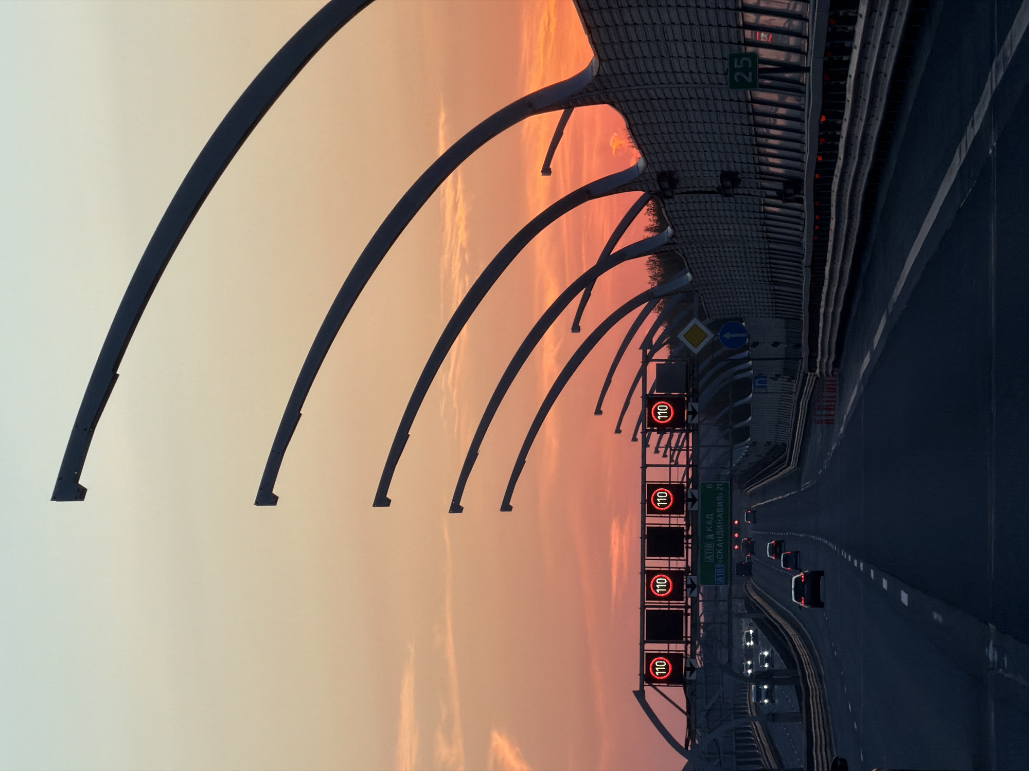 The image depicts the Western High-Speed Diameter (WHSD) highway in Saint Petersburg, Russia, at sunset. The view is of a multi-lane highway with several cars traveling on it. Overhead are a series of arches, part of the highway's structure, silhouetted against a pastel sky of pink, orange, and light yellow hues. To the side of the highway, there are speed limit signs displaying "110" within a red circle. Above the highway, a sign reads "25". Other road signs are also visible. The overall scene presents a modern infrastructure landscape against the backdrop of a colorful sunset.